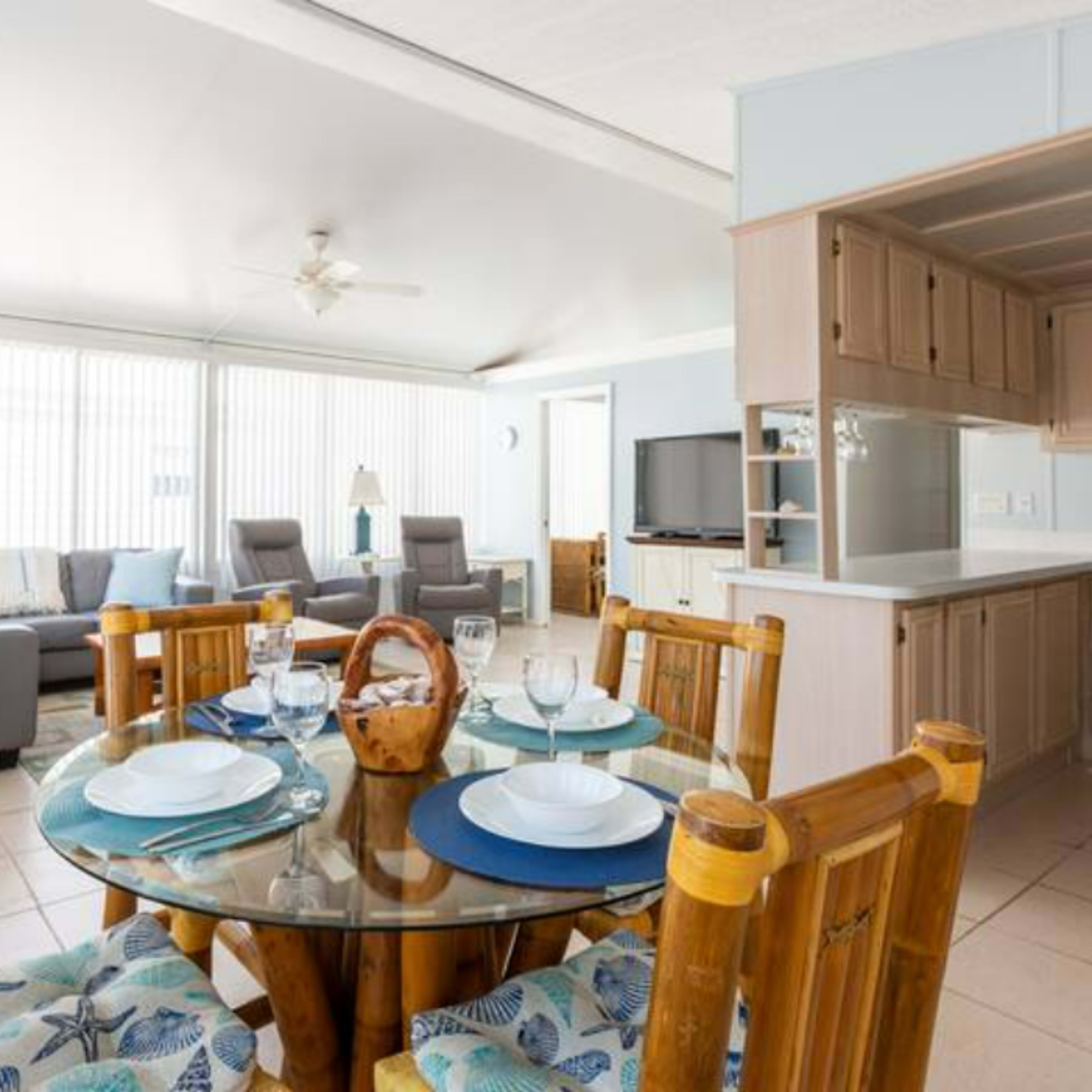 A bright open-concept living space featuring a dining area with a glass table and bamboo chairs, connected to a cozy sitting area with a couch and a television.