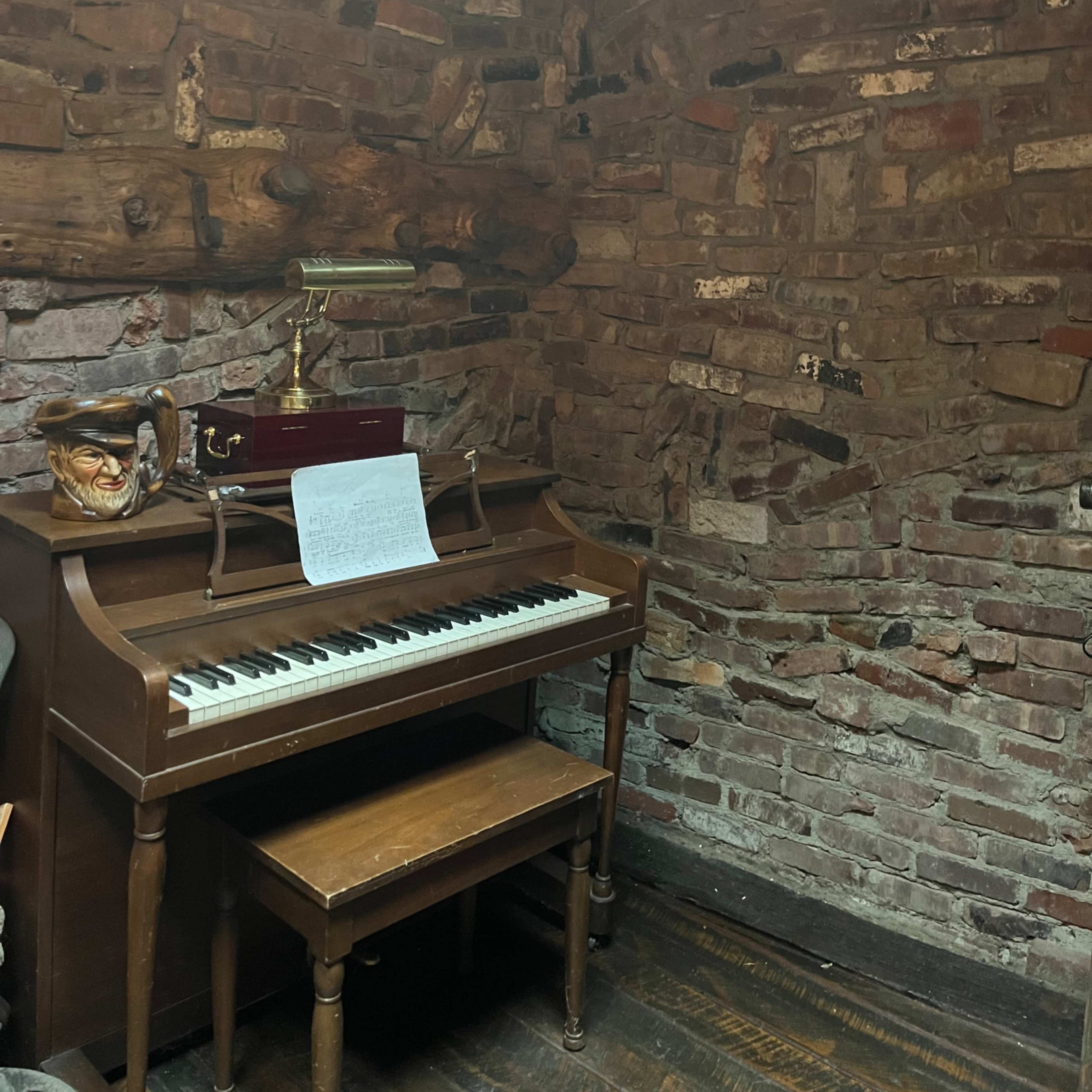 A wooden piano with sheet music sits against a brick wall in a dimly lit corner of a room.