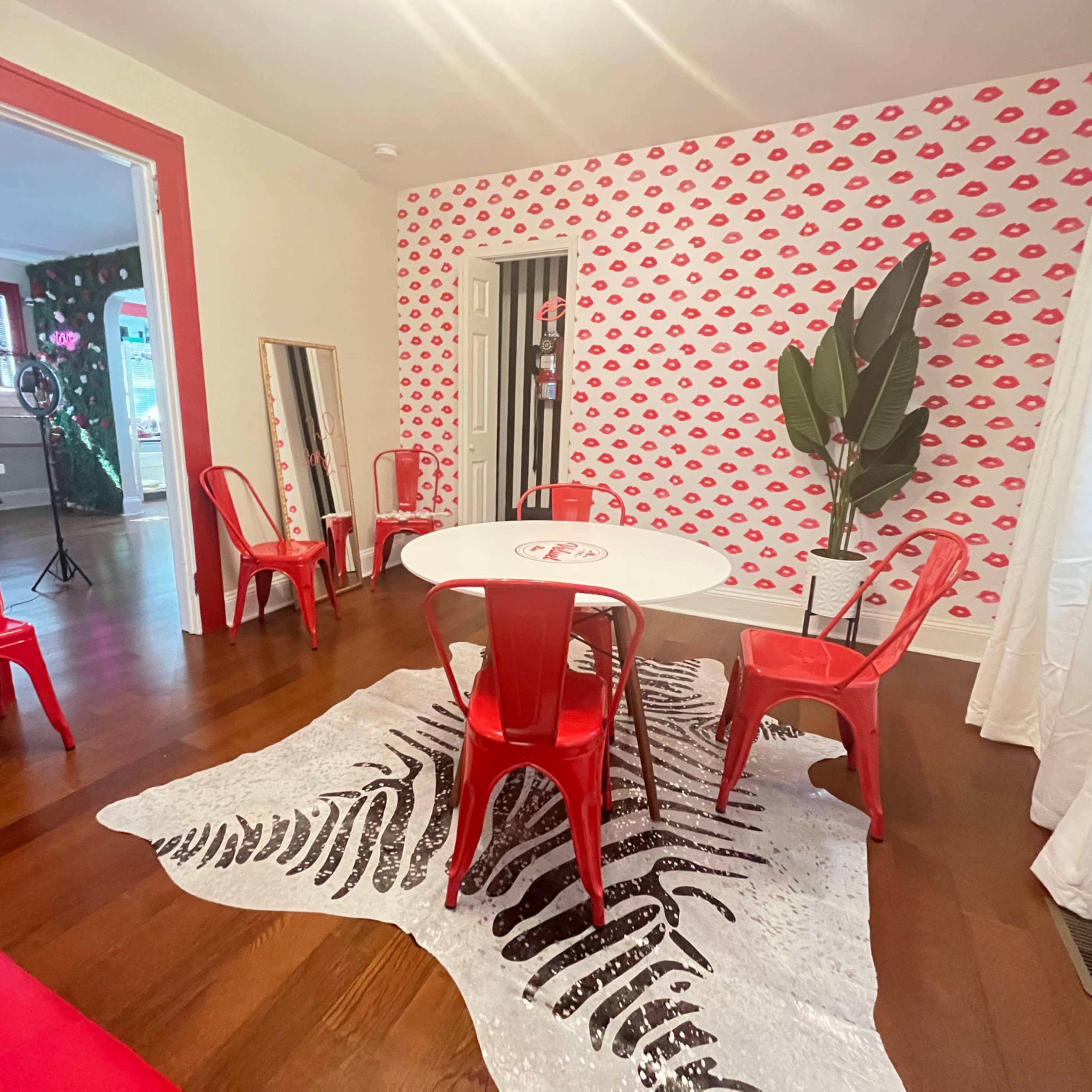 A modern room features a round white table surrounded by six red chairs, a patterned wall with lips designs, and a faux zebra rug on the hardwood floor.