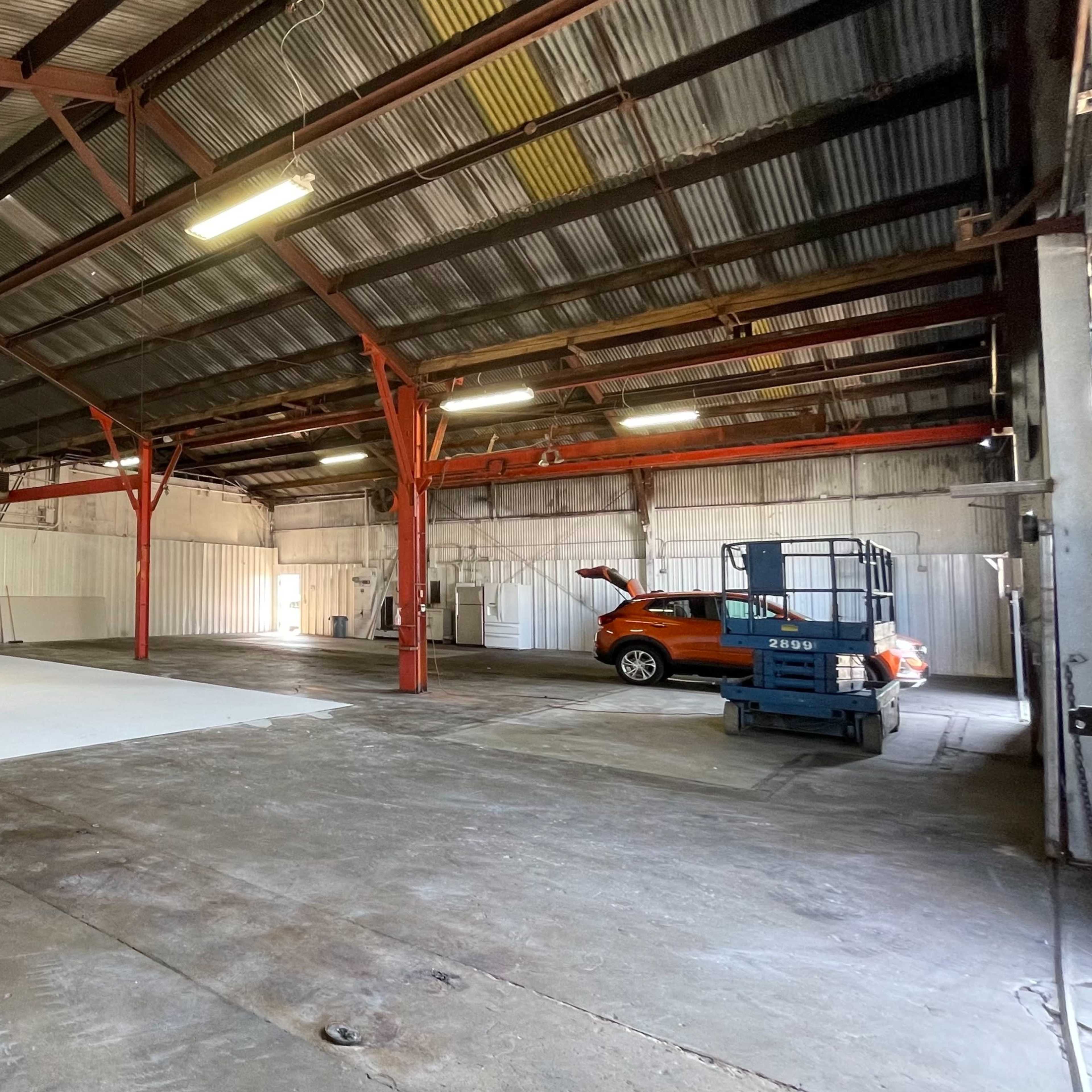 An industrial warehouse space with a large white backdrop and a cherry picker alongside a parked orange vehicle.