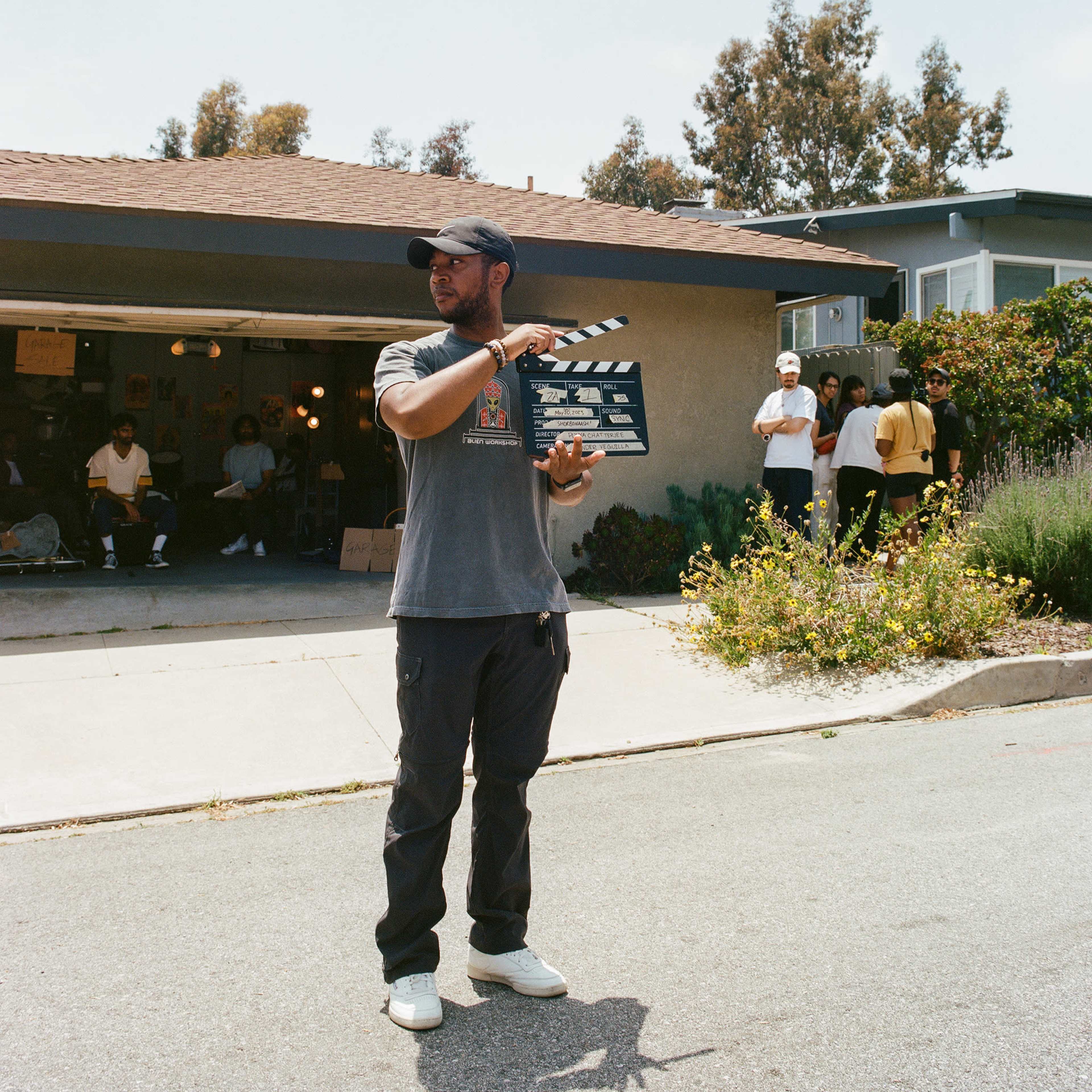 A person holds a movie clapperboard in front of a house while a group of people gathers in the background.