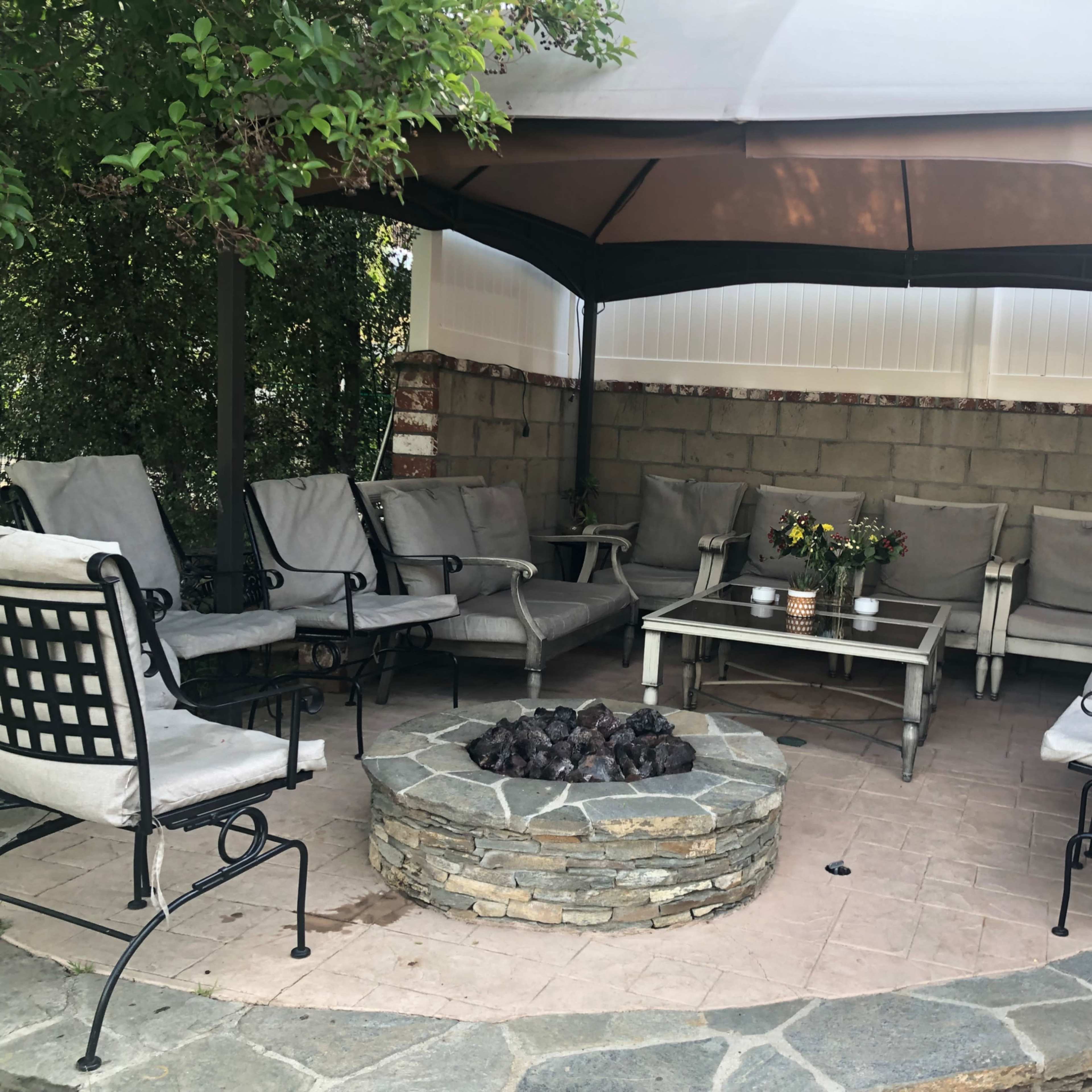 A covered outdoor seating area with a circular stone fire pit and multiple chairs arranged around a central table.