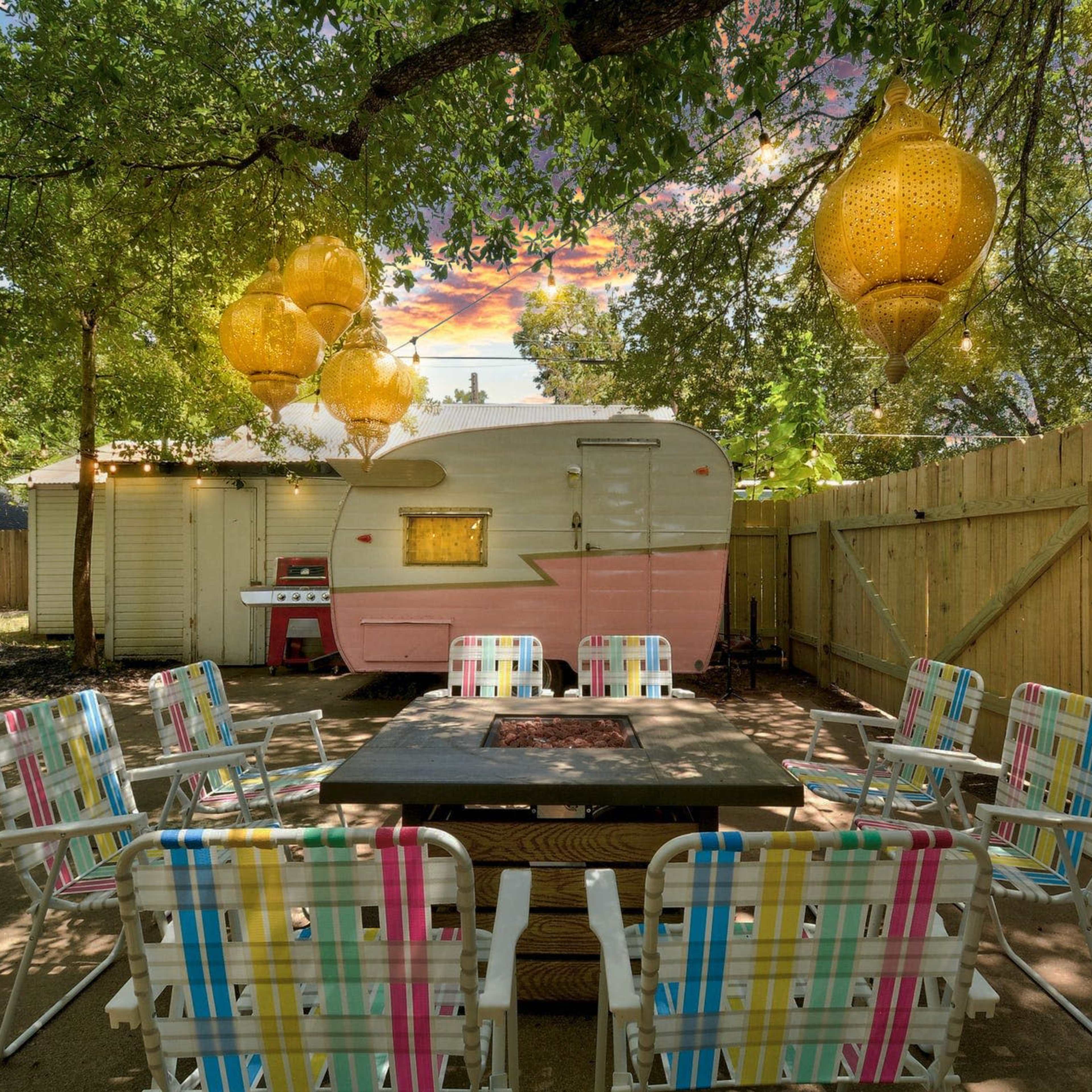 A colorful outdoor seating area with patterned chairs surrounds a rectangular fire pit, beside a vintage pink and white trailer and illuminated by lanterns hanging from tree branches.