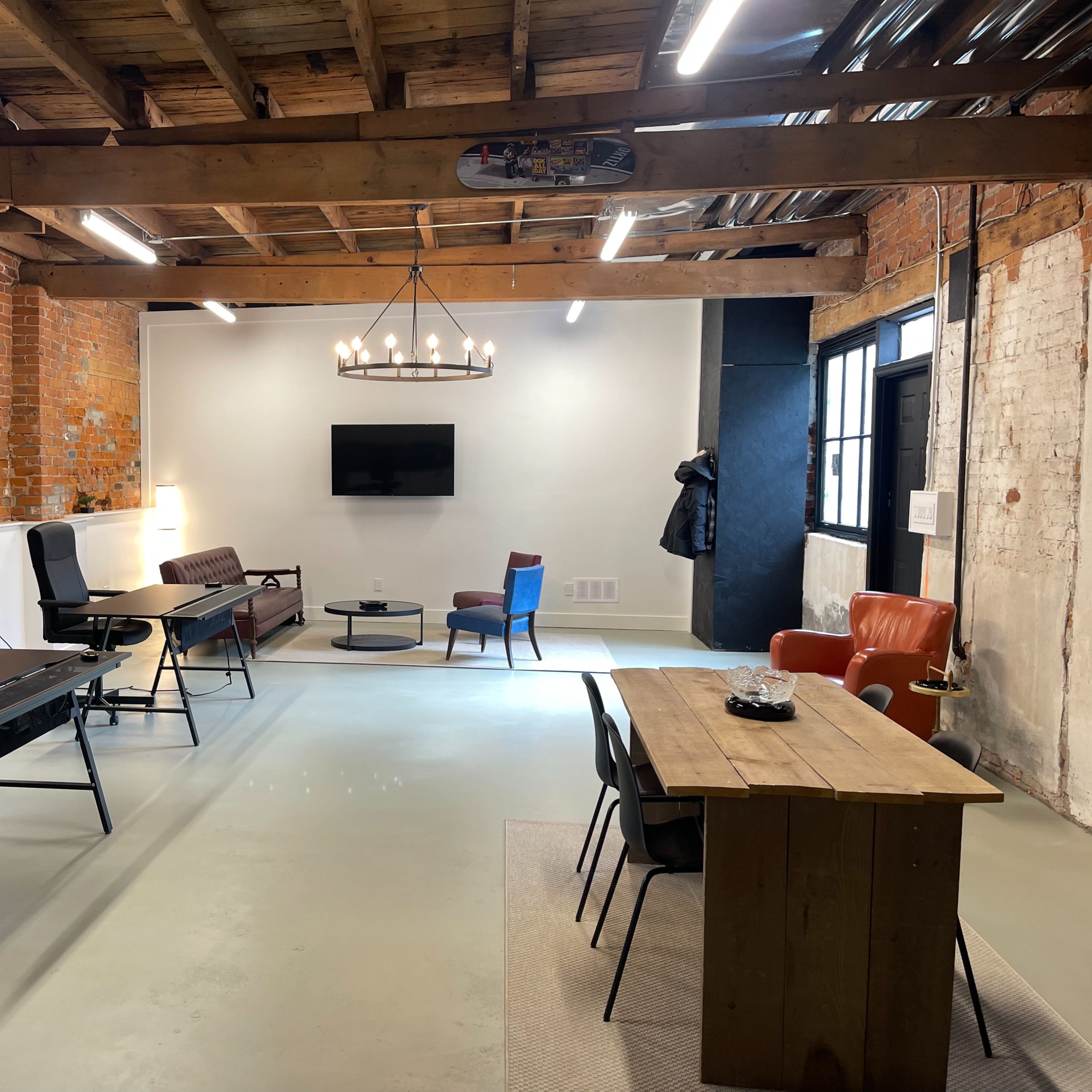 A spacious office interior features exposed brick walls, a wooden beam ceiling, a combination of modern and vintage furniture, and a large television mounted on the wall.