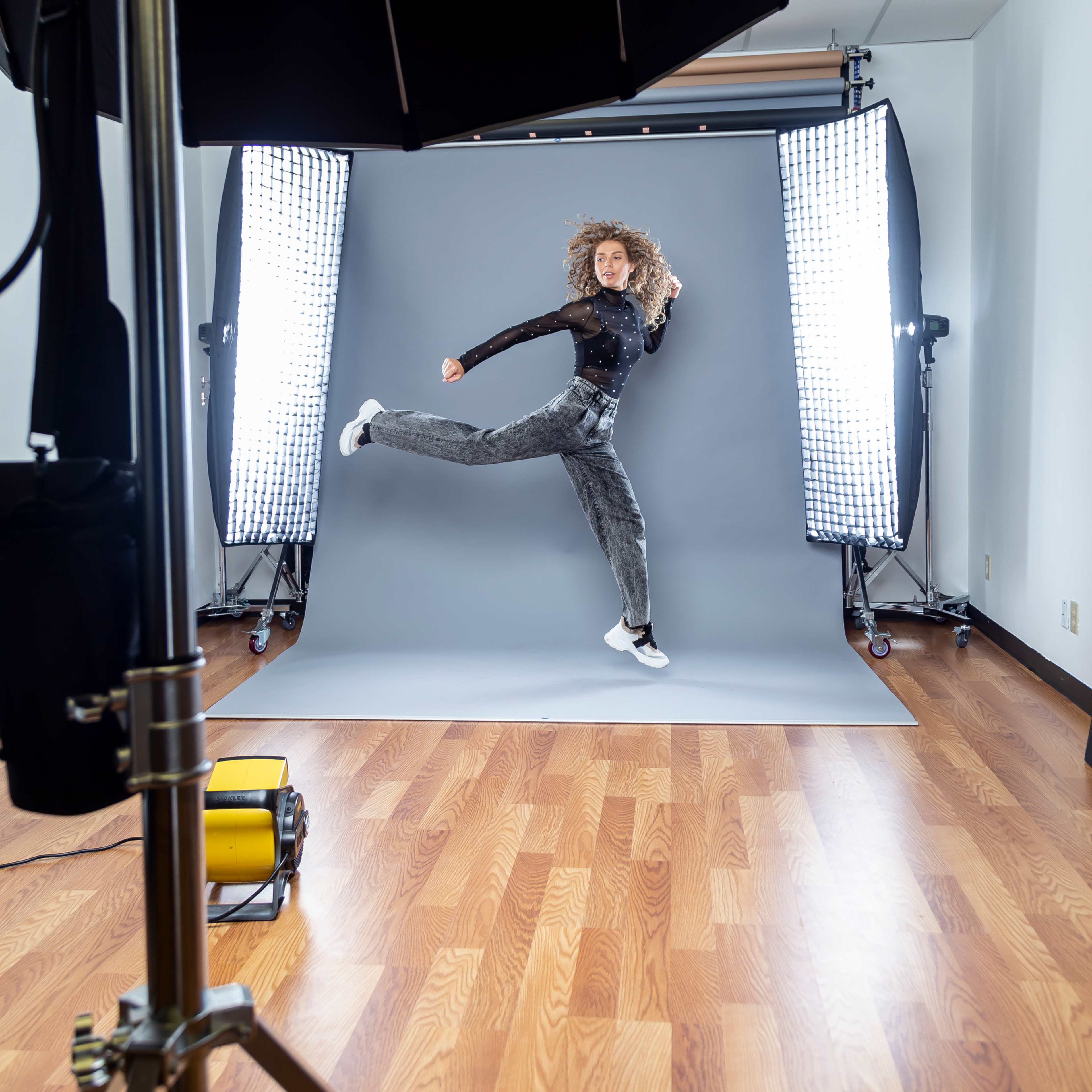 A person is jumping in a photography studio with lighting equipment and a gray backdrop.