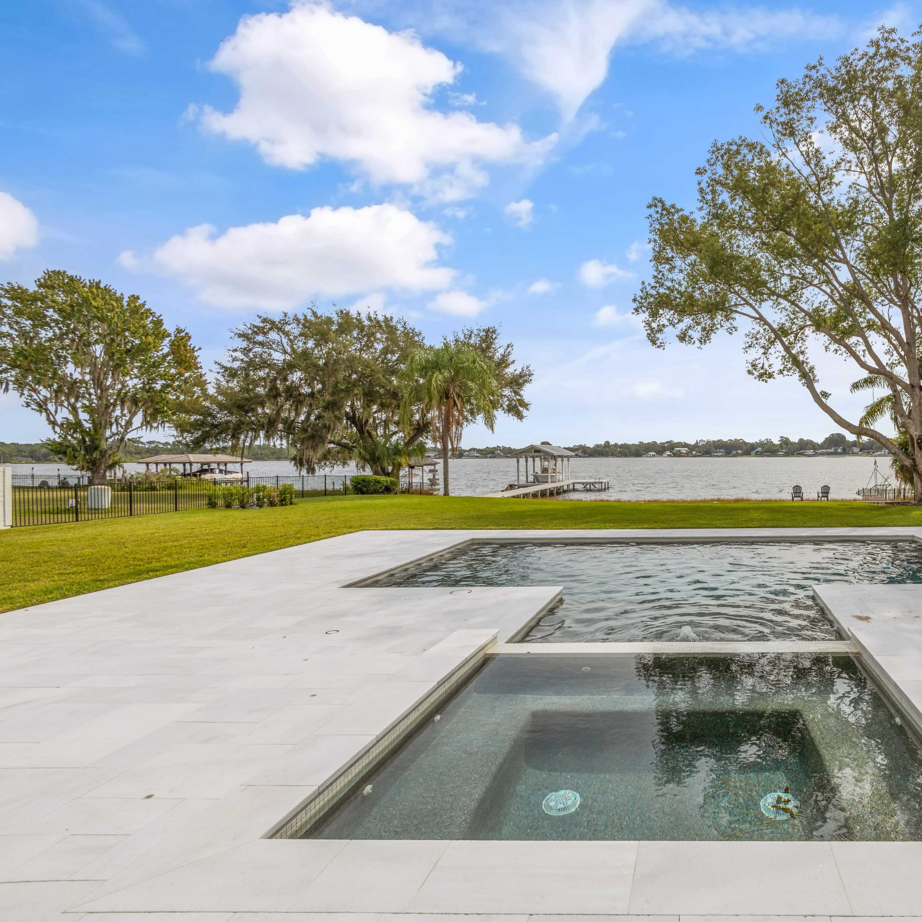 A modern outdoor area featuring a swimming pool and spa overlooking a calm body of water, surrounded by green grass and trees.