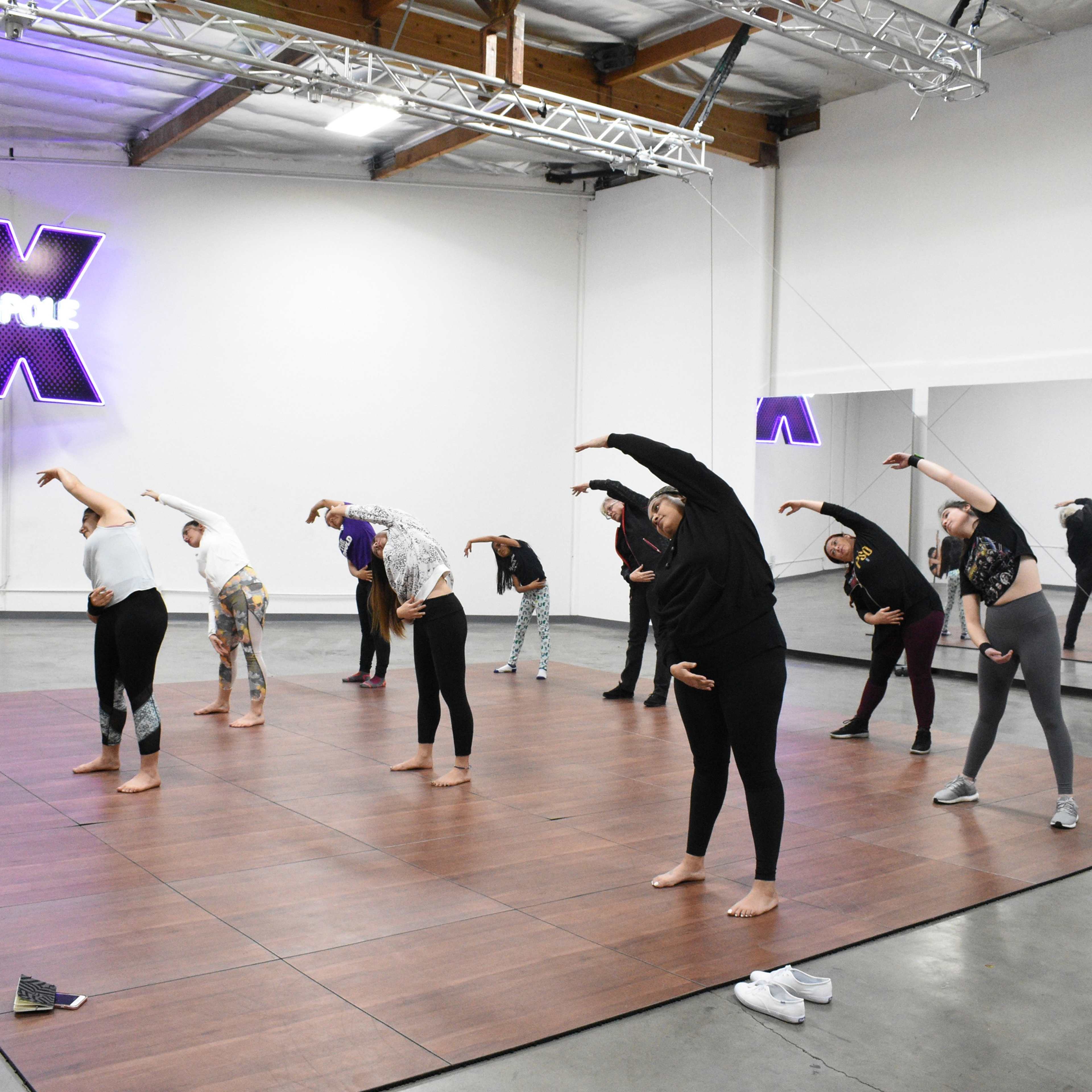 A group of people is practicing stretching exercises on a wooden dance floor in a studio with neon lighting.