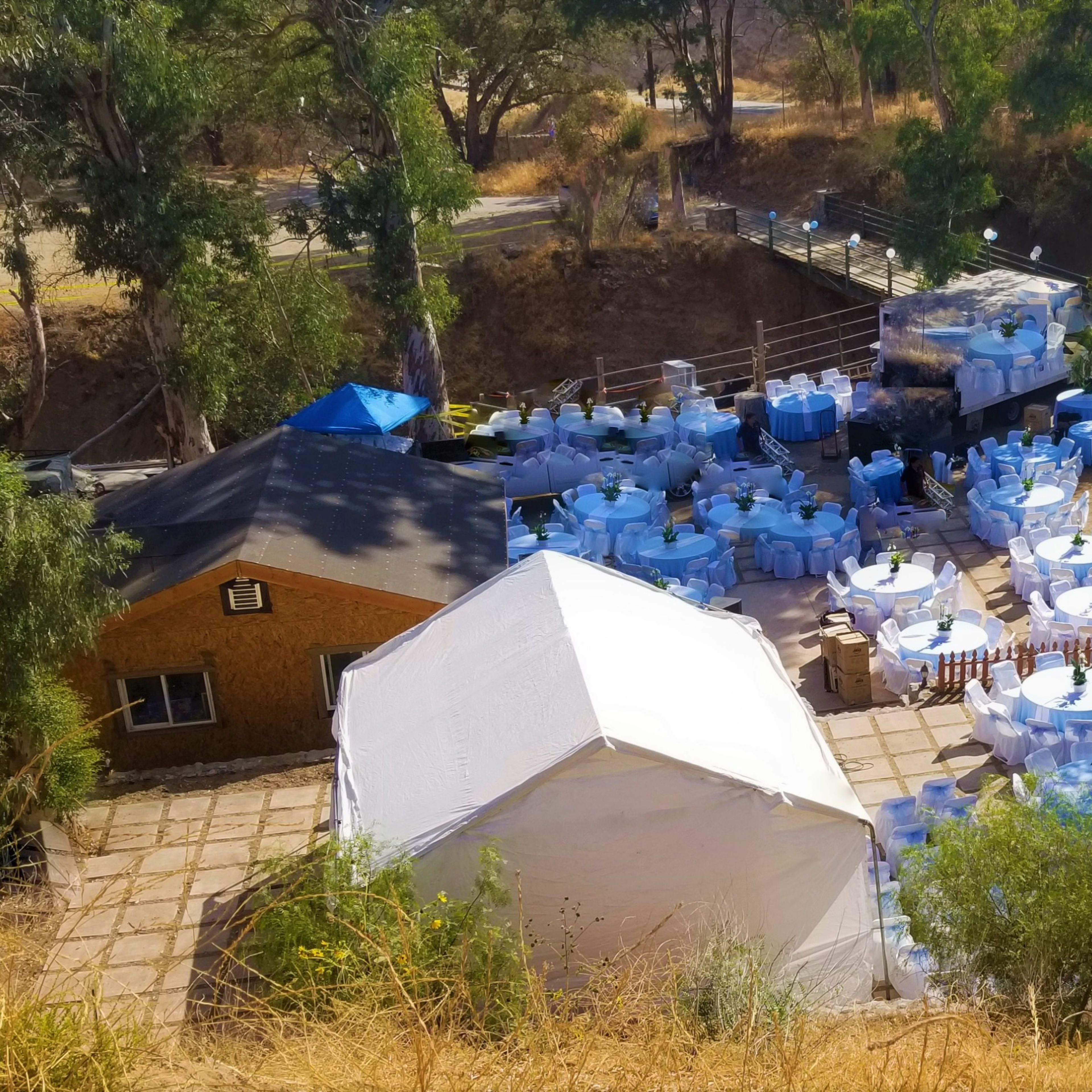 An outdoor event setup with numerous round tables covered in light blue tablecloths surrounded by greenery.