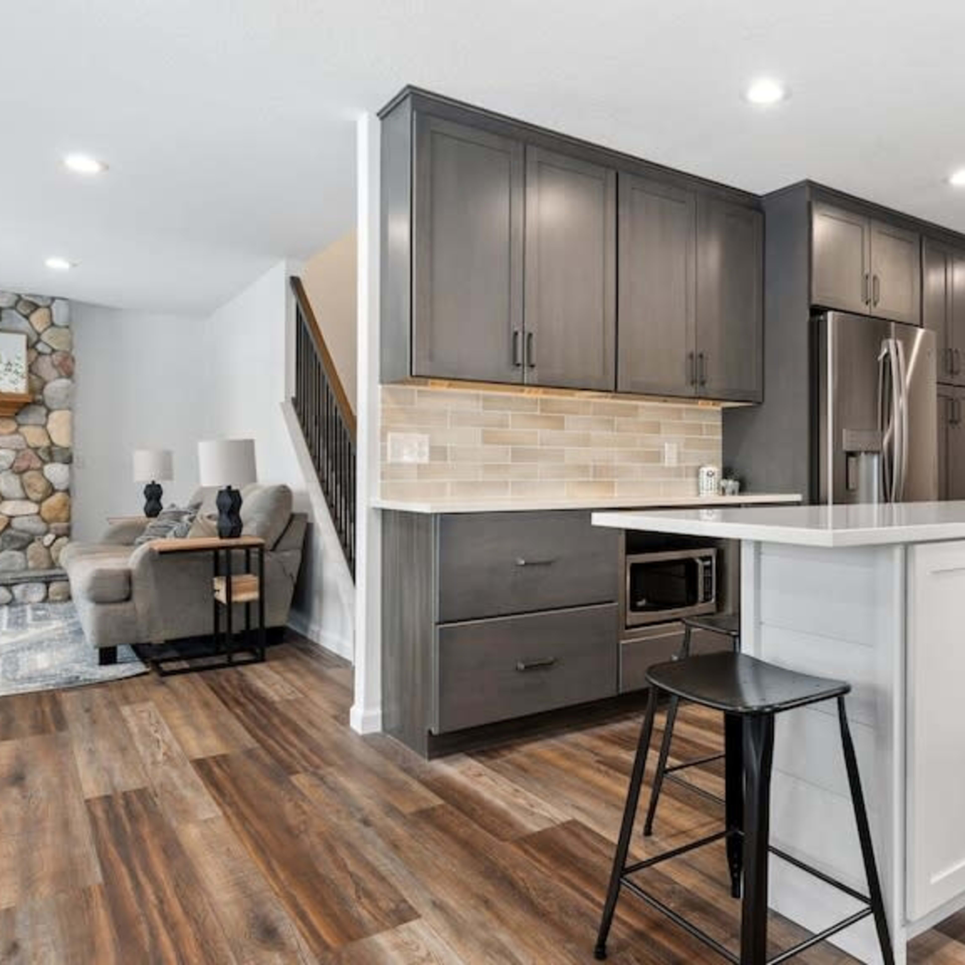 A modern kitchen with dark cabinetry, a stainless steel refrigerator, and an island with a black stool, connected to a living area featuring a stone fireplace.