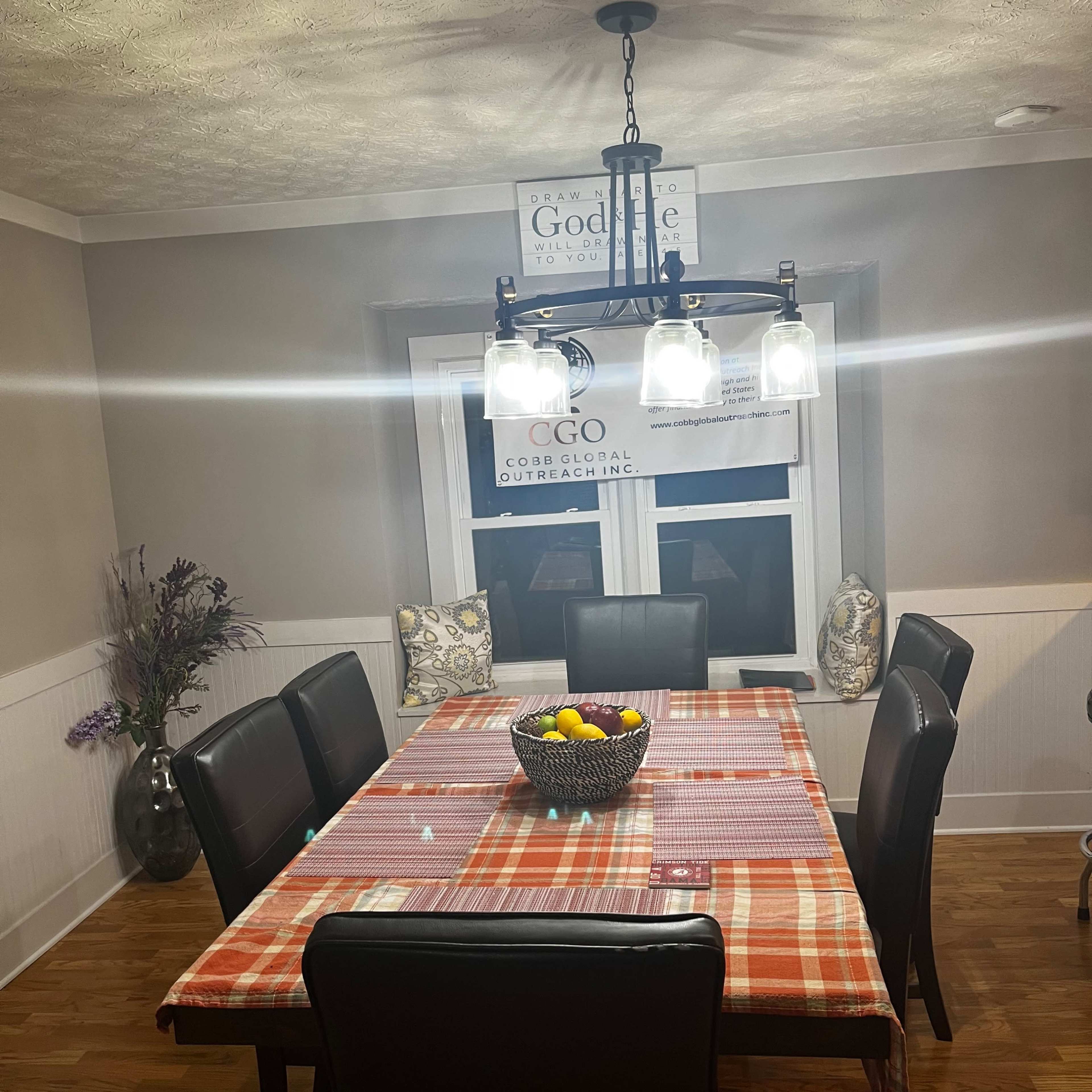 A spacious dining room features a long table set with a checkered tablecloth, surrounded by black chairs and a chandelier above, with a fruit bowl in the center.