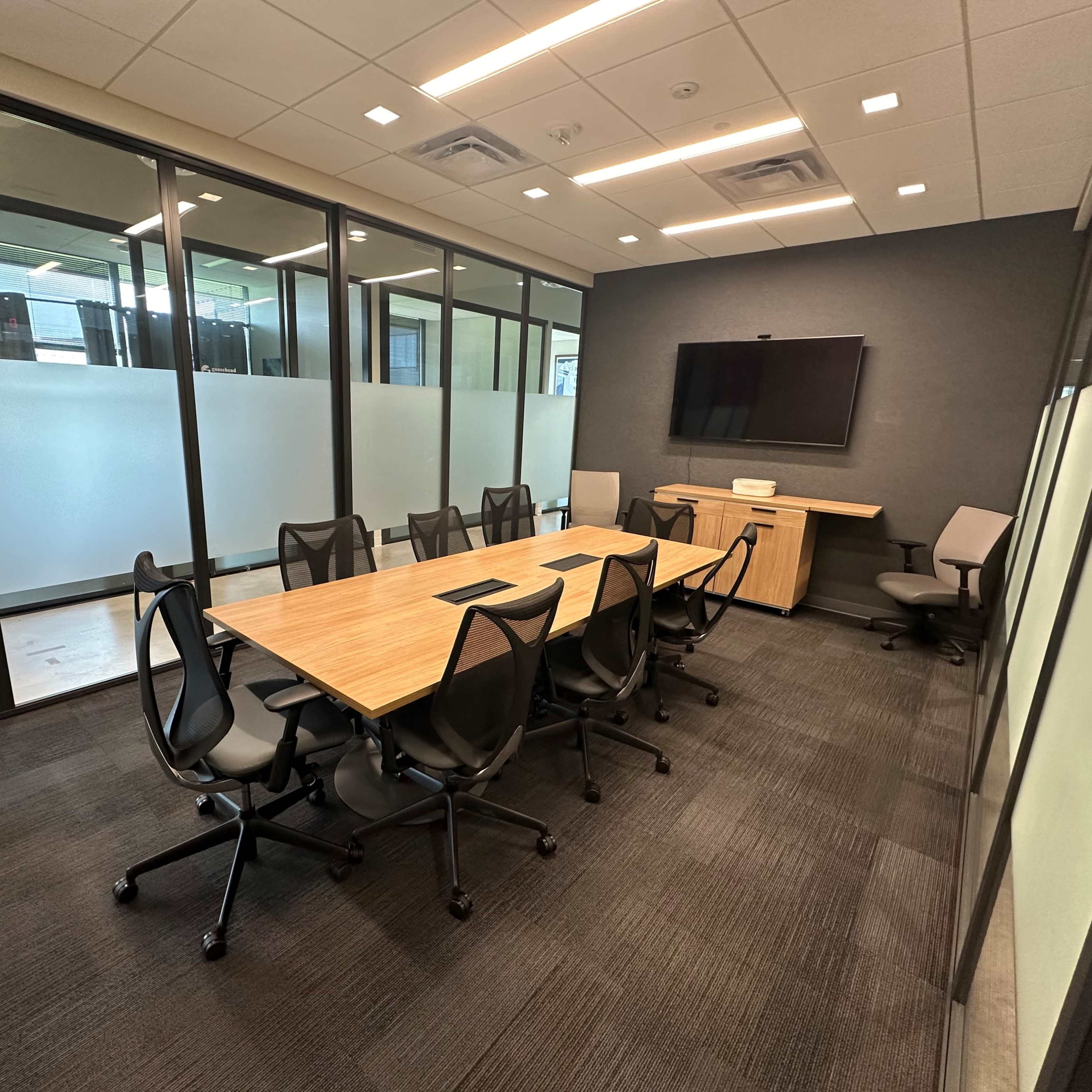 A modern conference room with a wooden table, several black chairs, and a wall-mounted television.