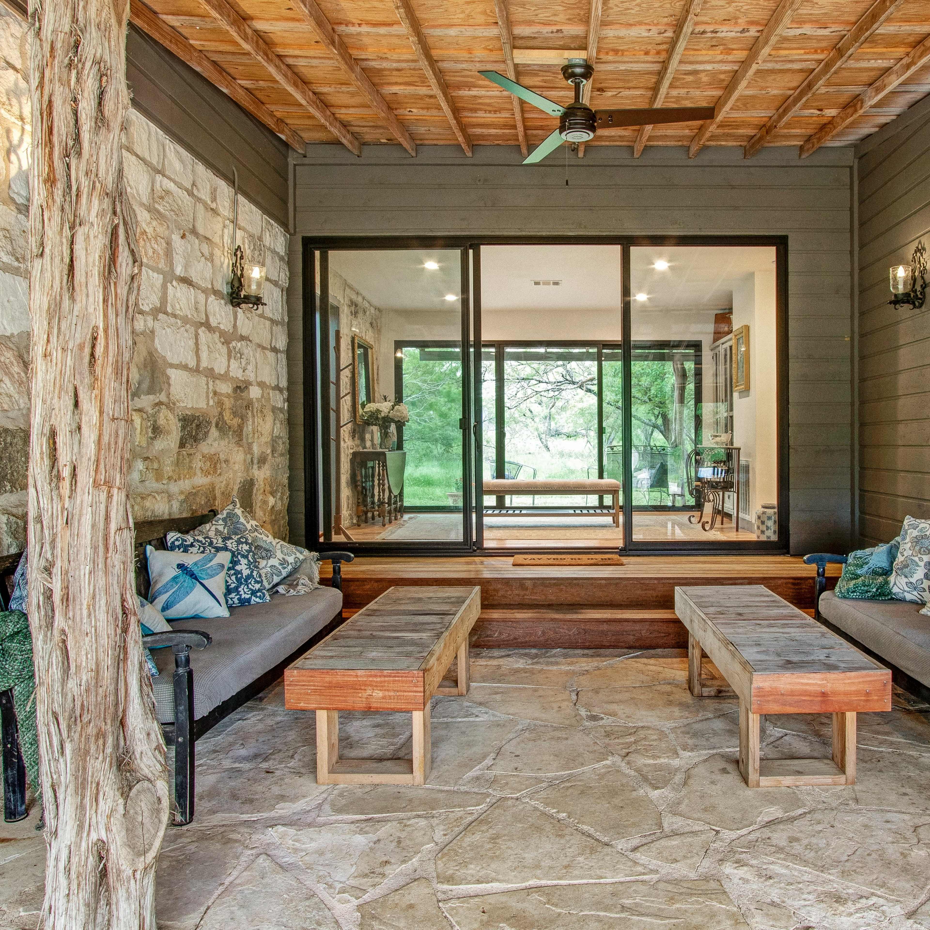 A covered porch features stone and wooden walls, two long wooden benches, and large glass doors opening to a view of greenery.