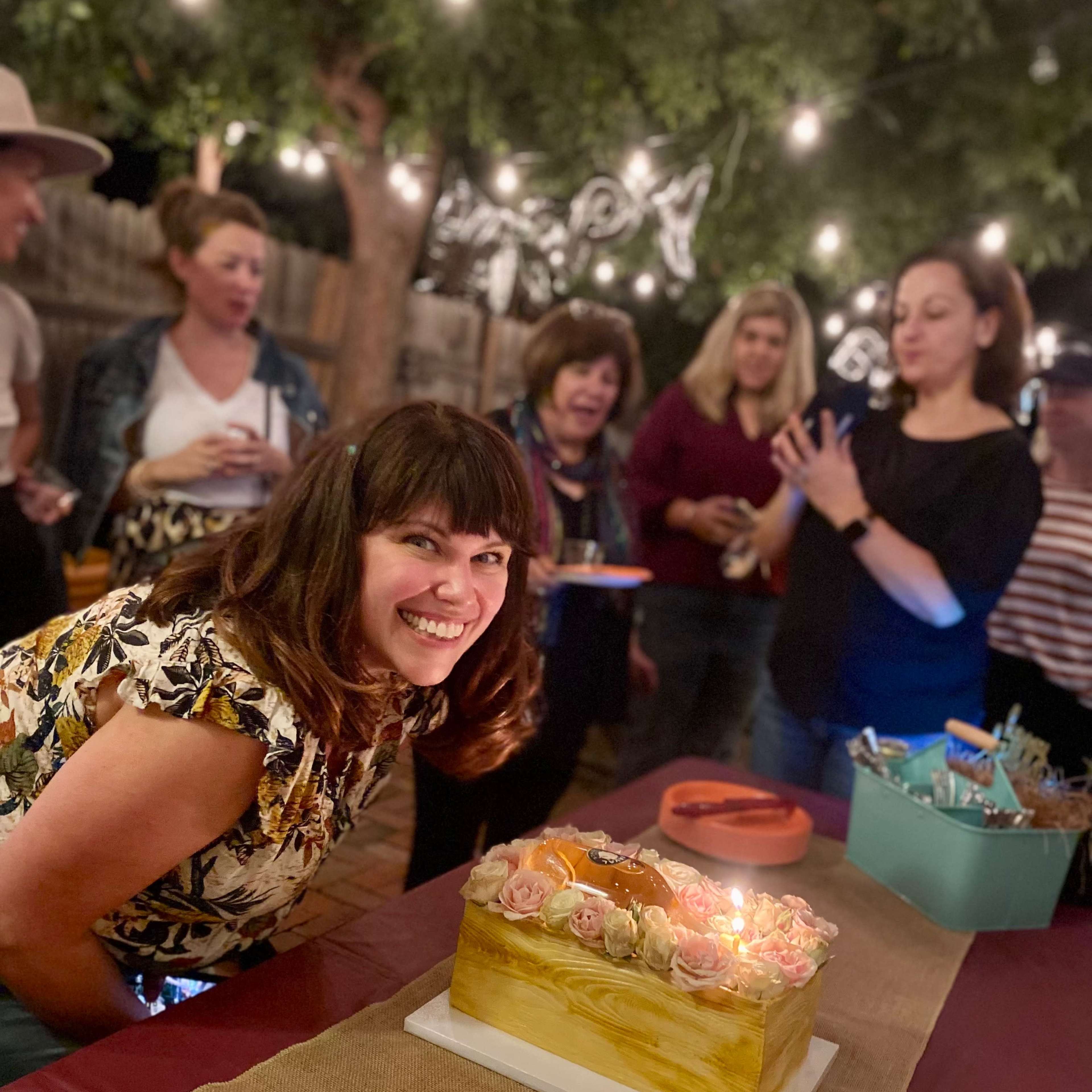 A woman smiles joyfully next to a decorated cake while a group of people celebrate in a warmly lit outdoor setting.