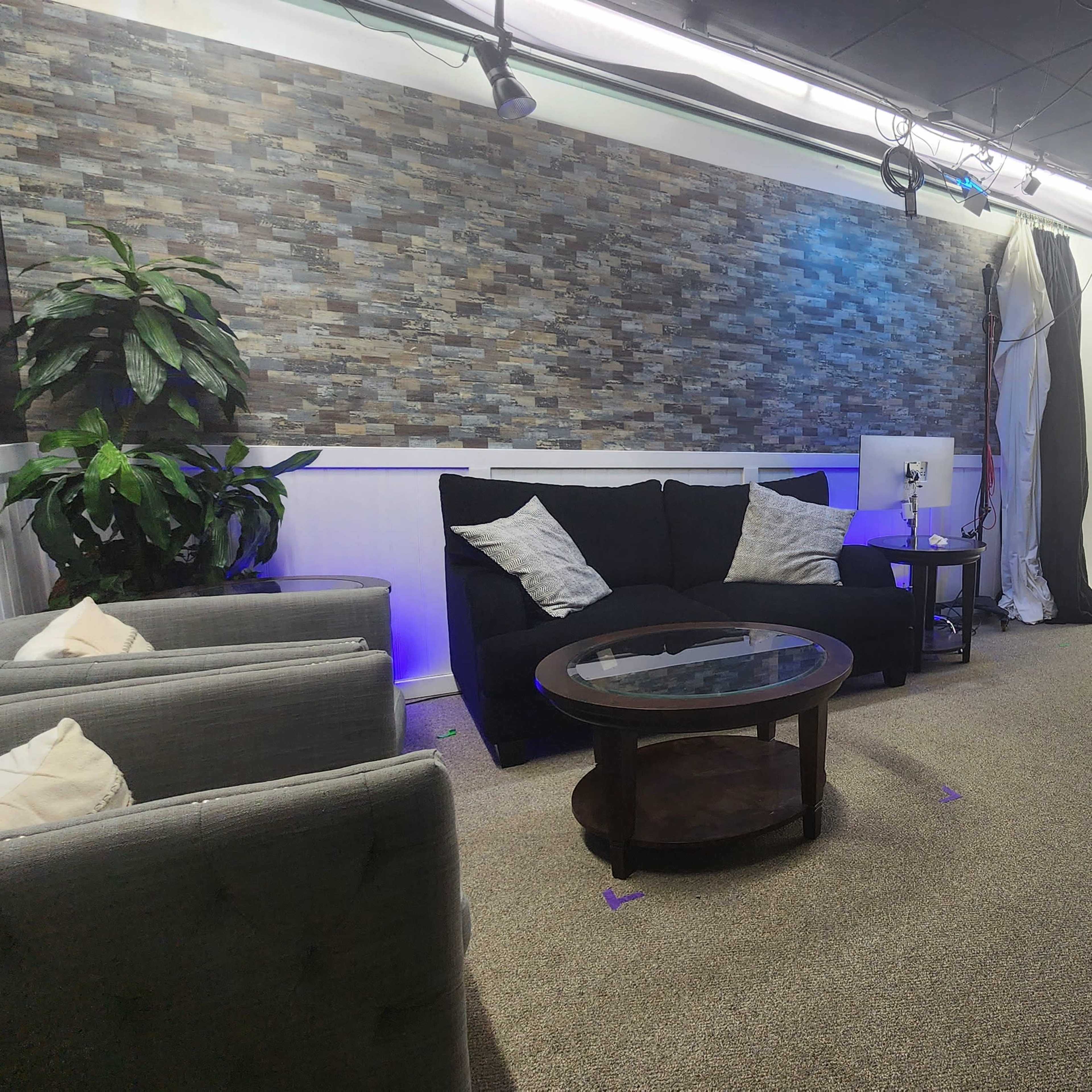 A modern studio setup with a textured stone wall, a black sofa with decorative pillows, a glass coffee table, and a large empty white backdrop.