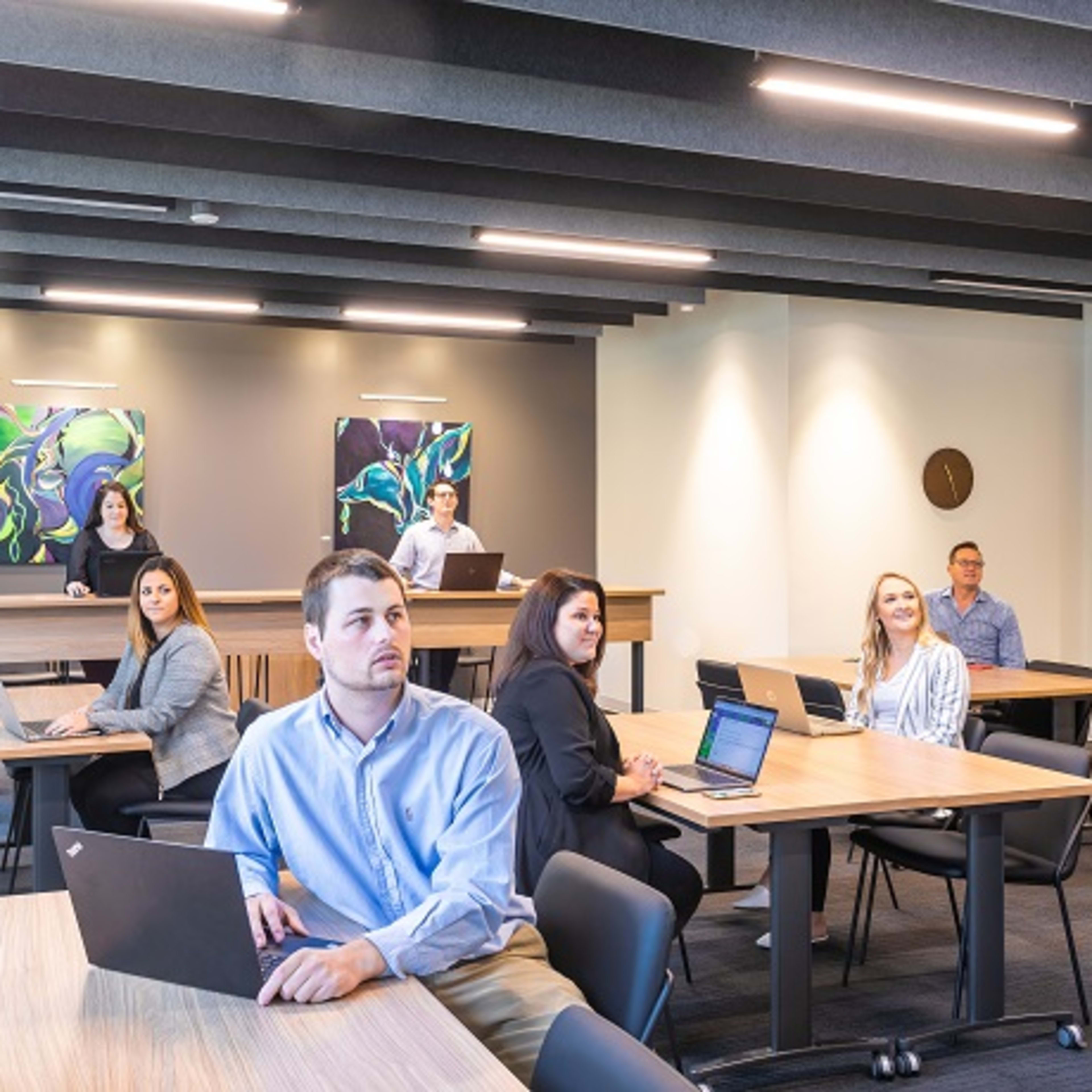 A group of eight professionals sits at tables in a modern conference room, focused on laptops with artwork displayed on the walls.