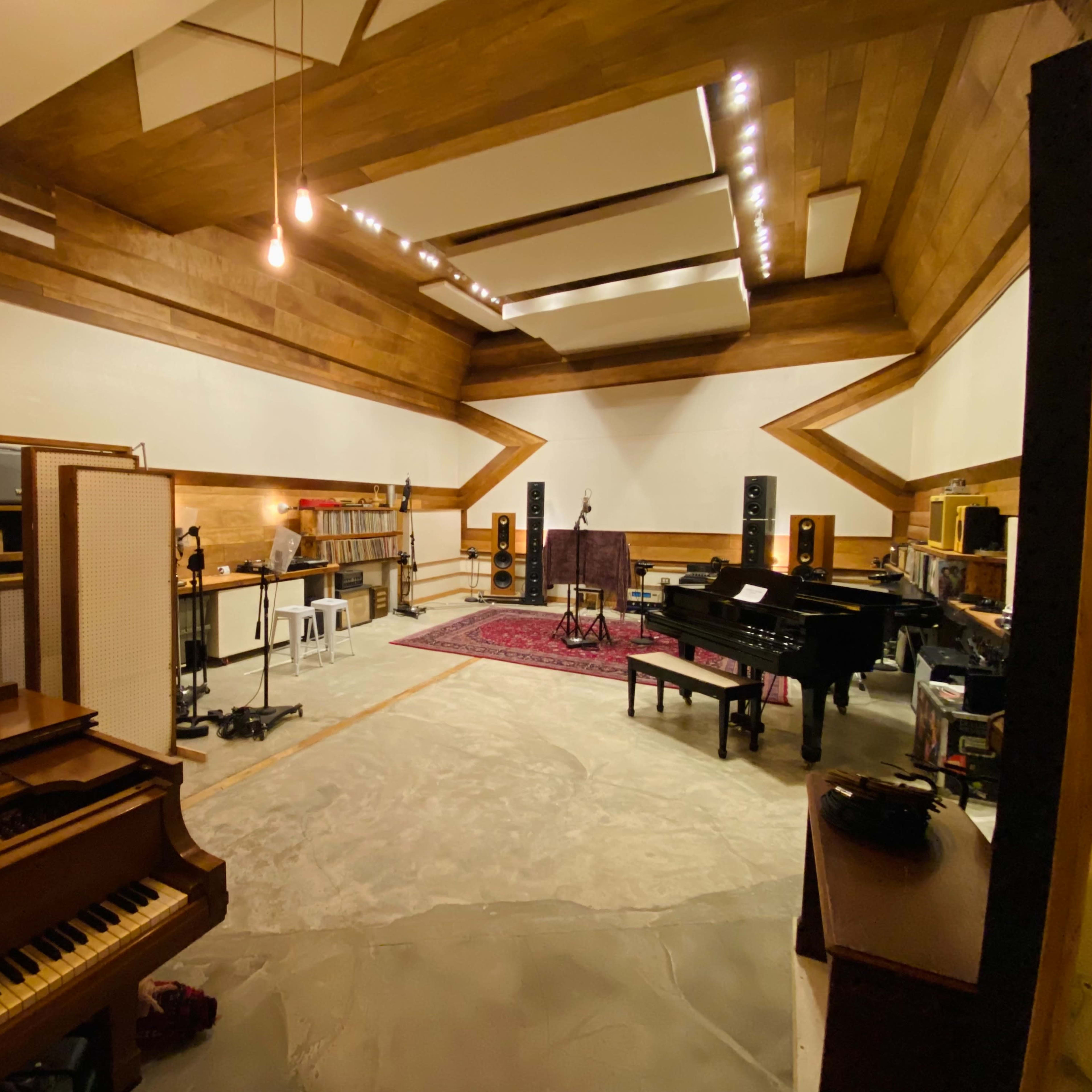 A spacious music studio with wooden paneling, a grand piano, and various audio equipment arranged around a central recording area.
