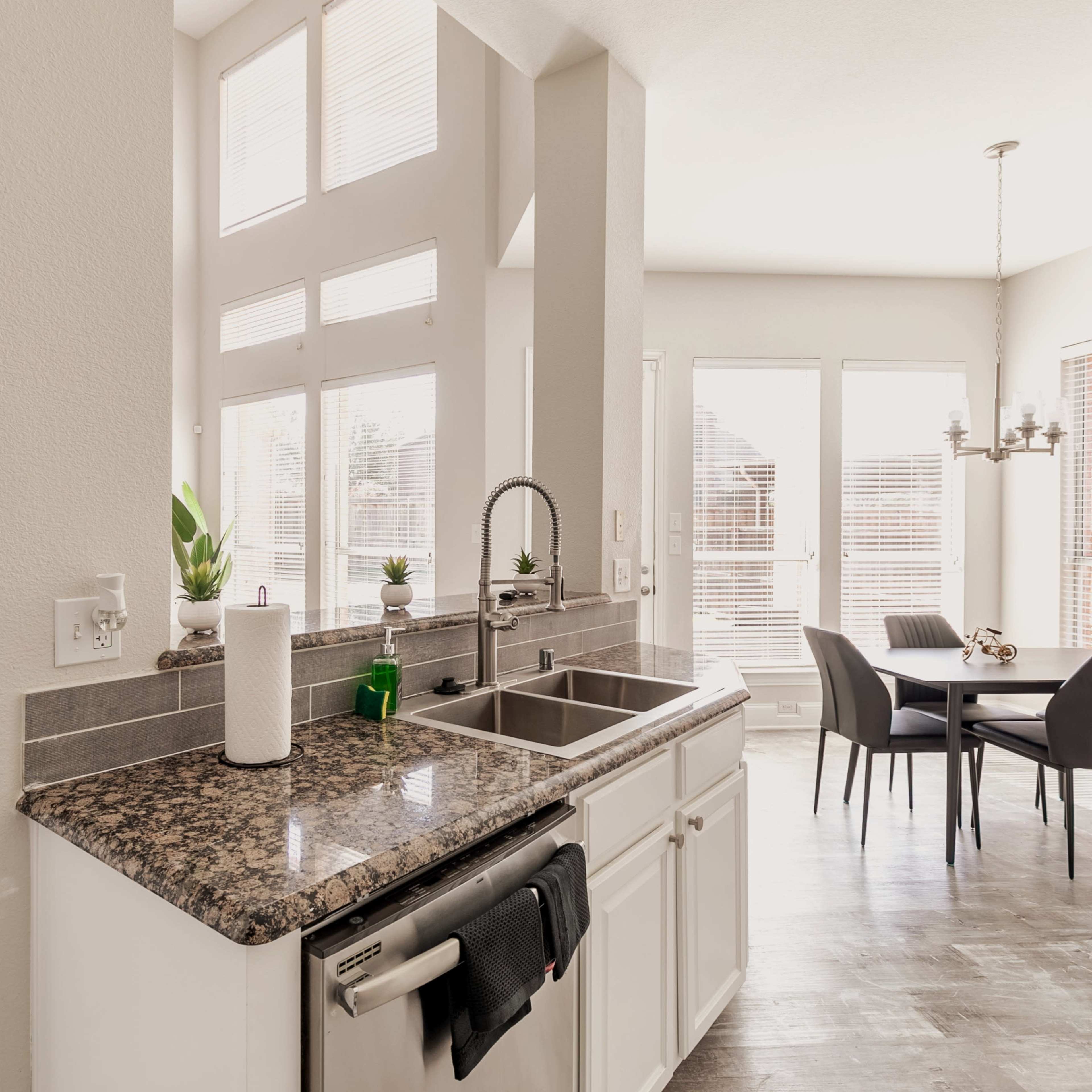 A modern kitchen with granite countertops, a stainless steel sink, and a dining area featuring a round table and chairs, illuminated by natural light from large windows.
