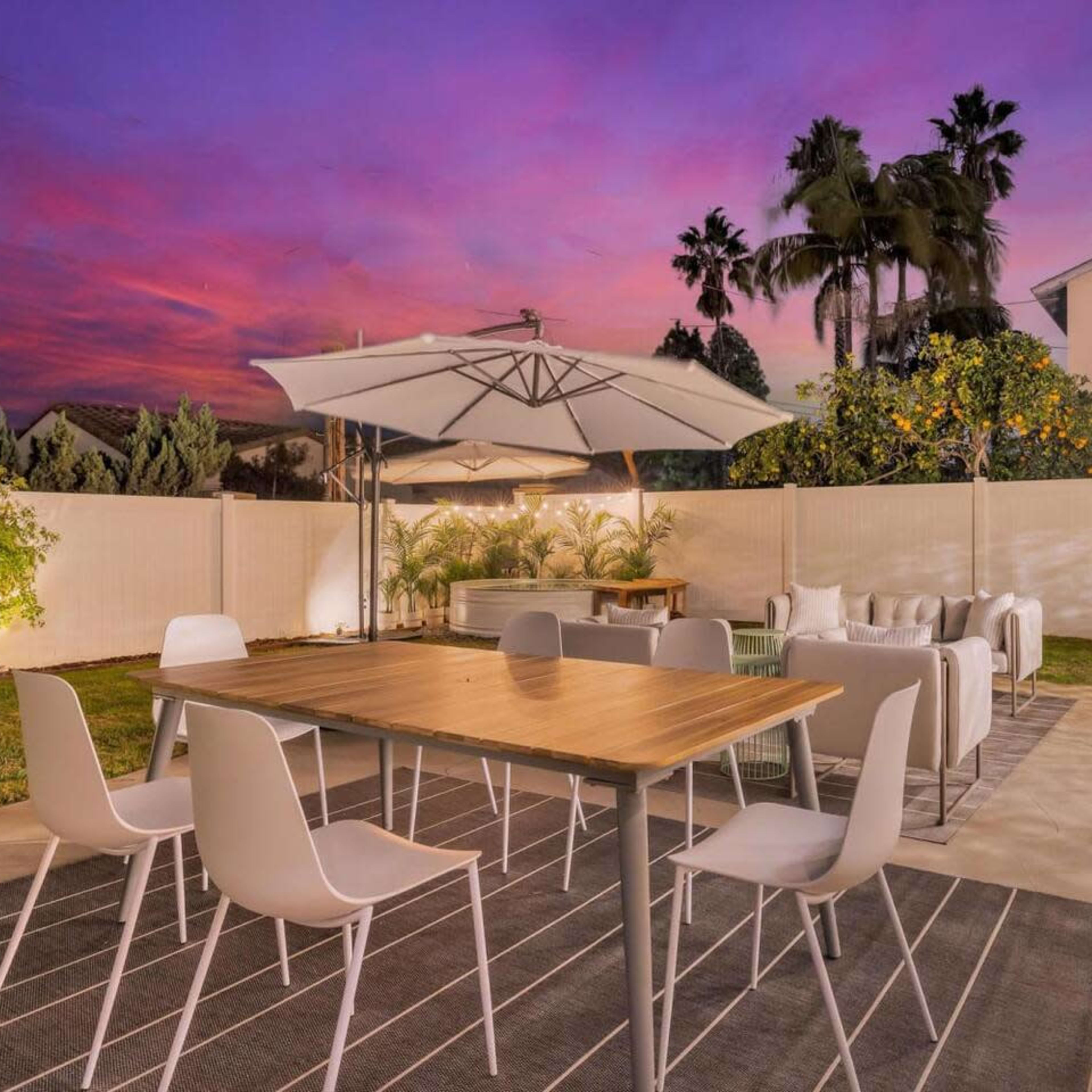 A backyard with a wooden dining table surrounded by white chairs, an umbrella overhead, and an outdoor seating area, set against a colorful sunset sky.