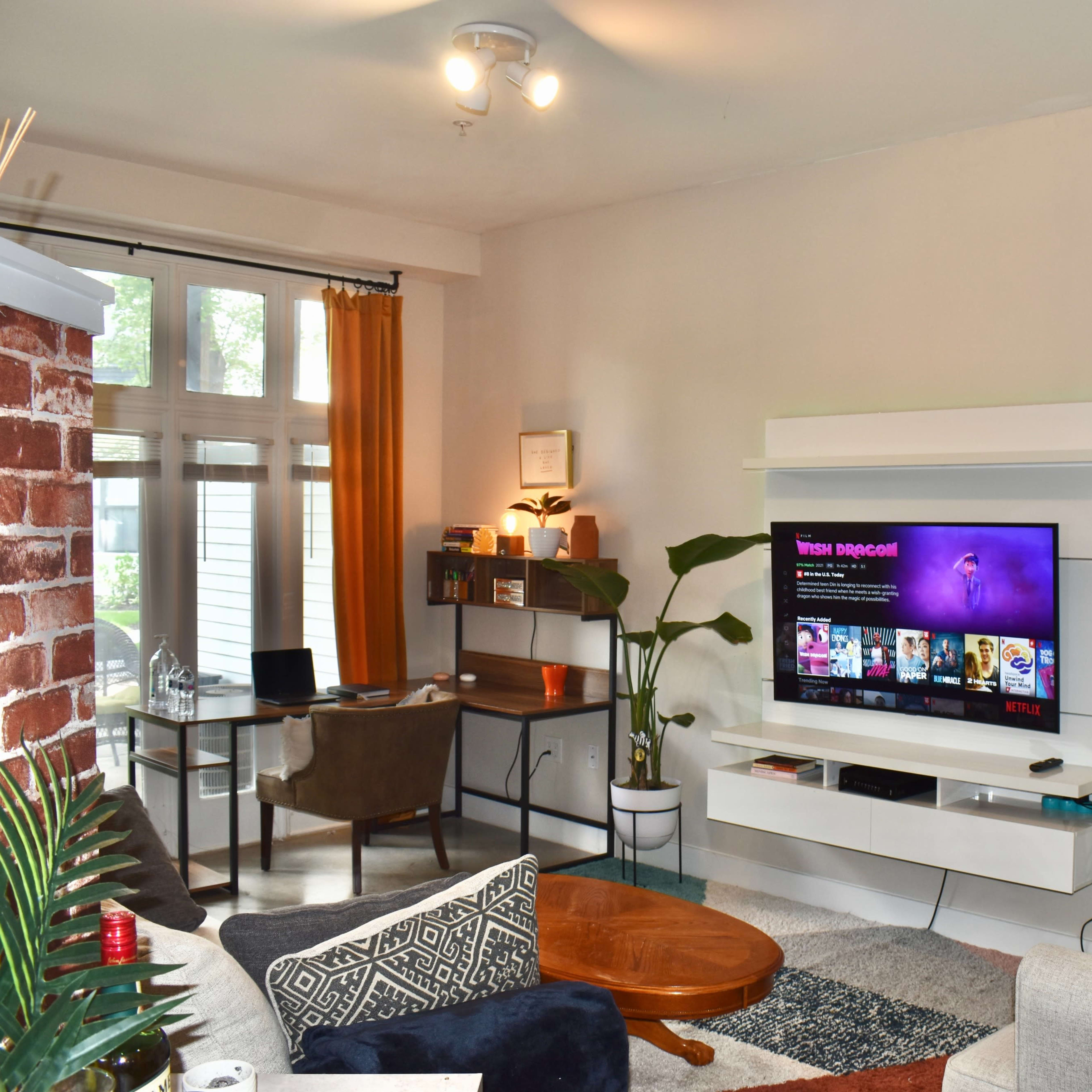 A living room features a mounted television displaying vibrant content, a cozy sofa, a circular coffee table, and a desk setup against a backdrop of large windows.