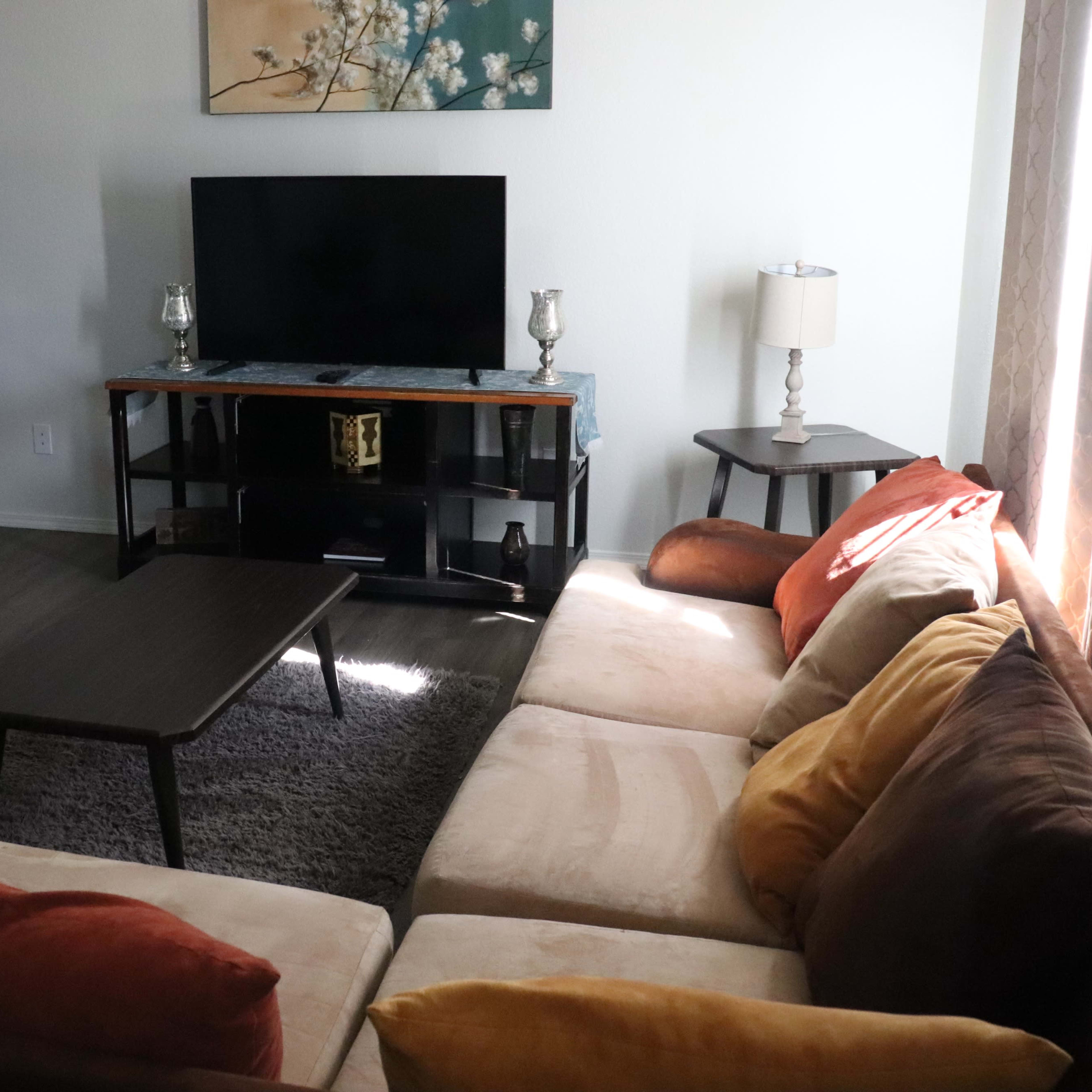 A living room features a sectional sofa, a television on a stand, and a small coffee table, with a doorway leading to another room in the background.