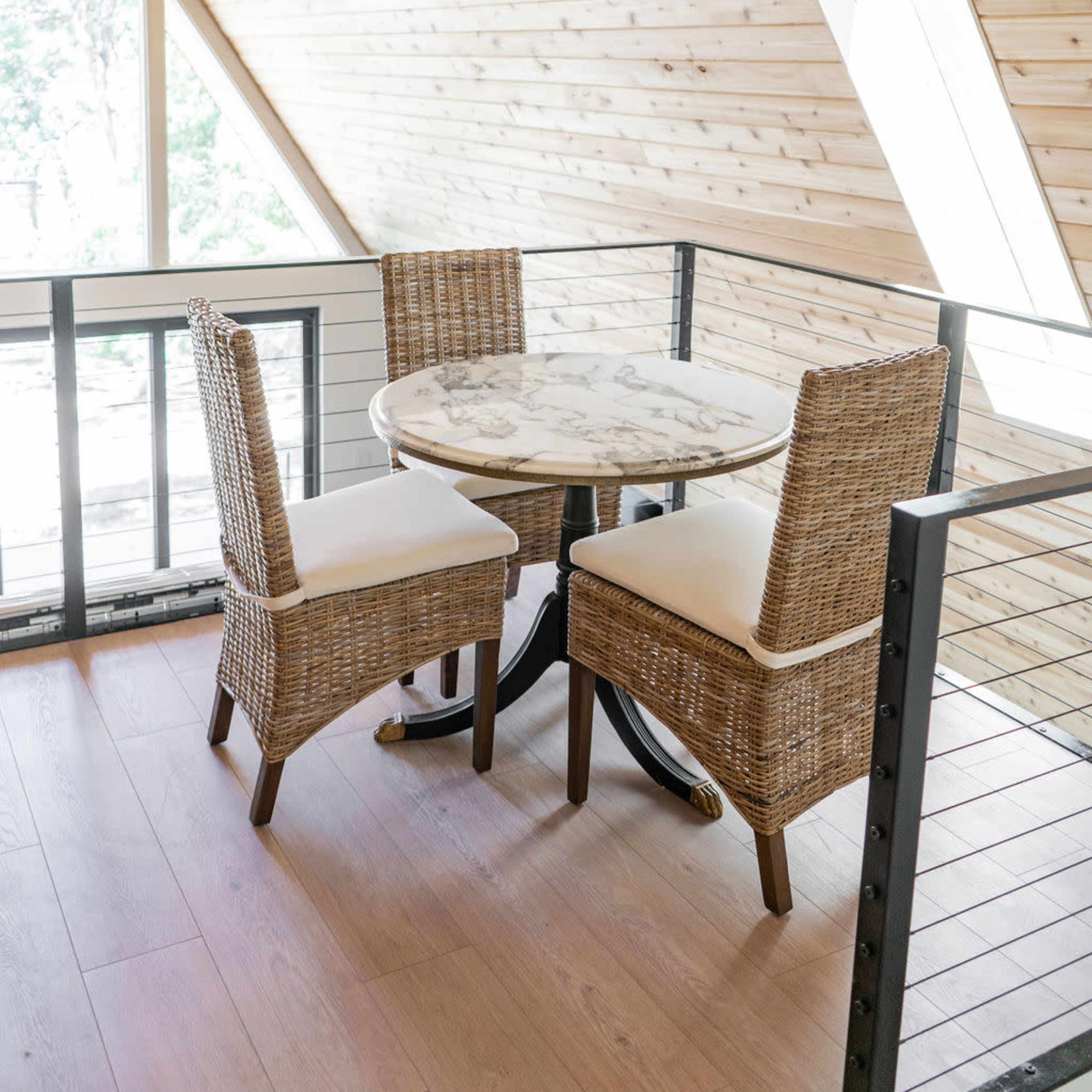 A round marble table with two wicker chairs is situated next to a window in a wooden interior space.