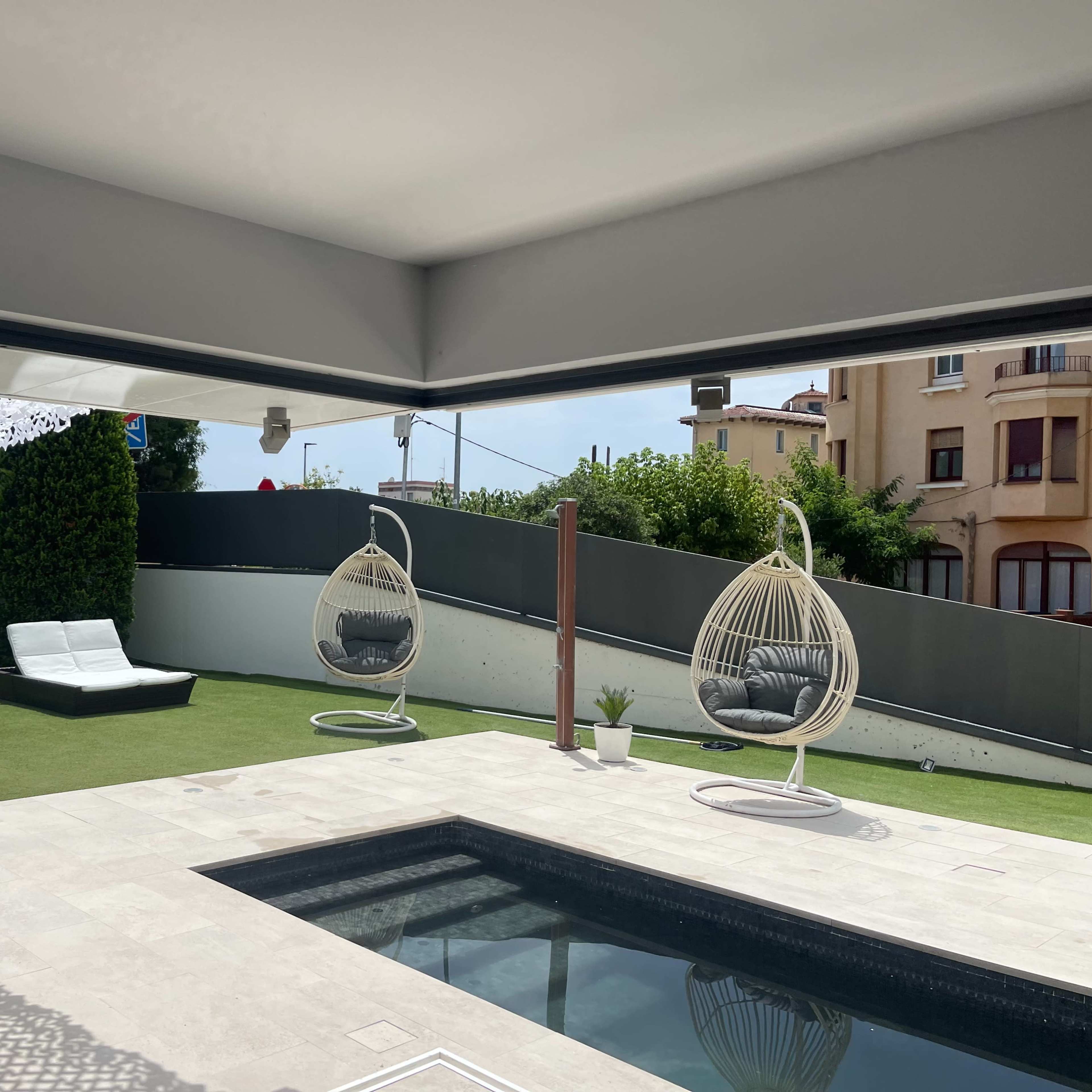 A modern outdoor patio features a pool, two hanging chairs, a sun lounger, and a neatly maintained lawn.