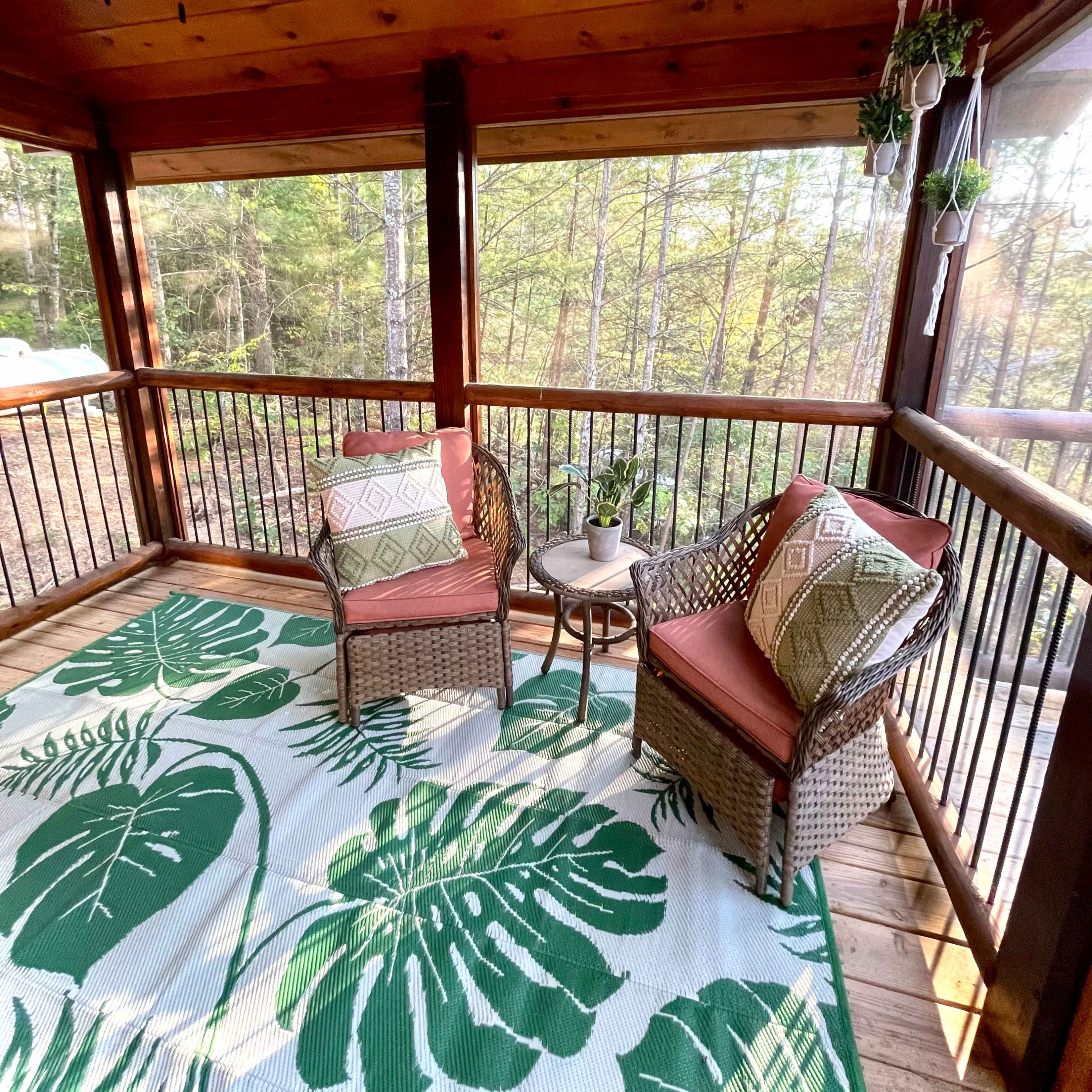 A screened-in porch features two wicker chairs with cushions, a small table, and a large green leaf-patterned rug, surrounded by trees.