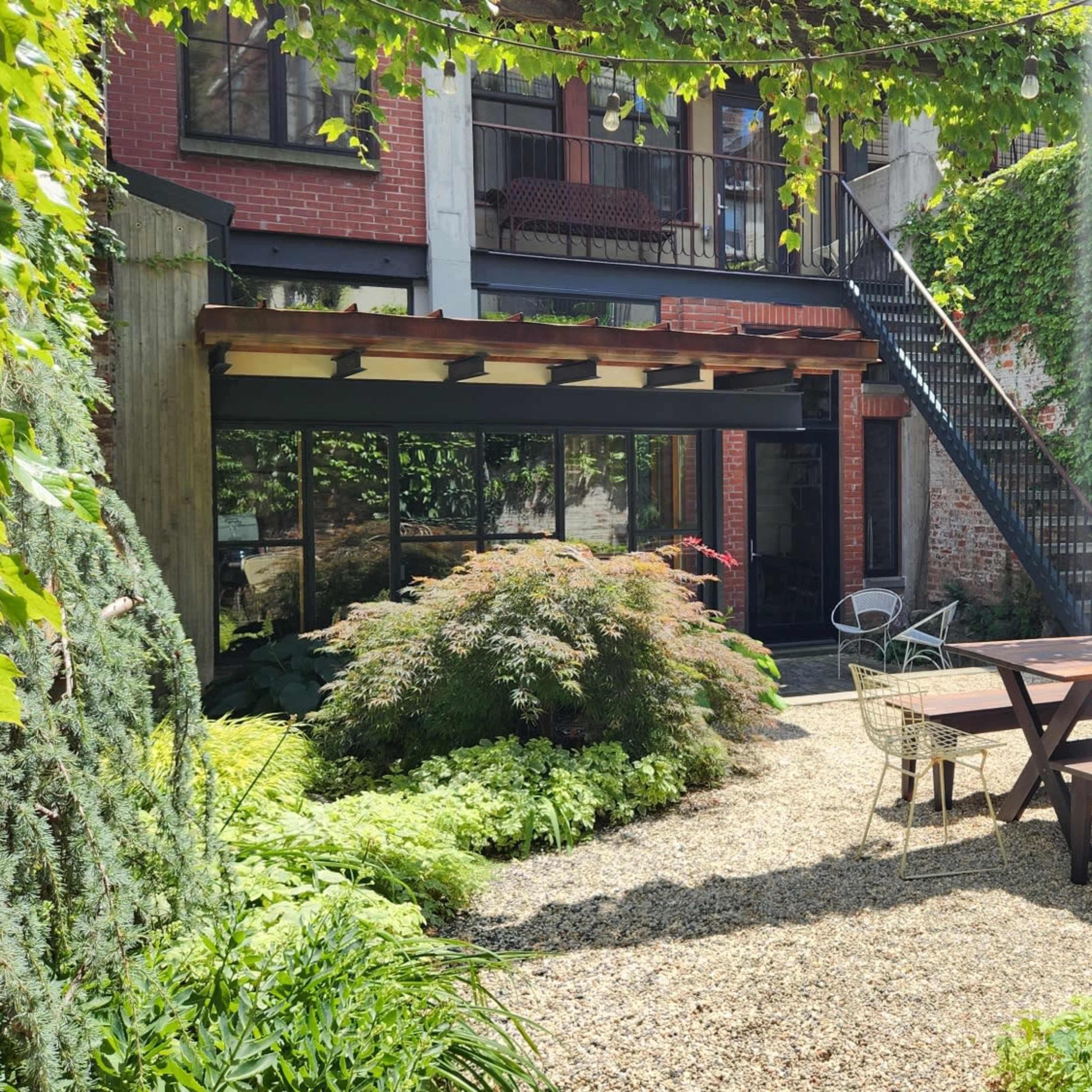 A sunlit courtyard with a wooden picnic table surrounded by lush greenery, stone gravel, and a building featuring stairs and balconies in the background.