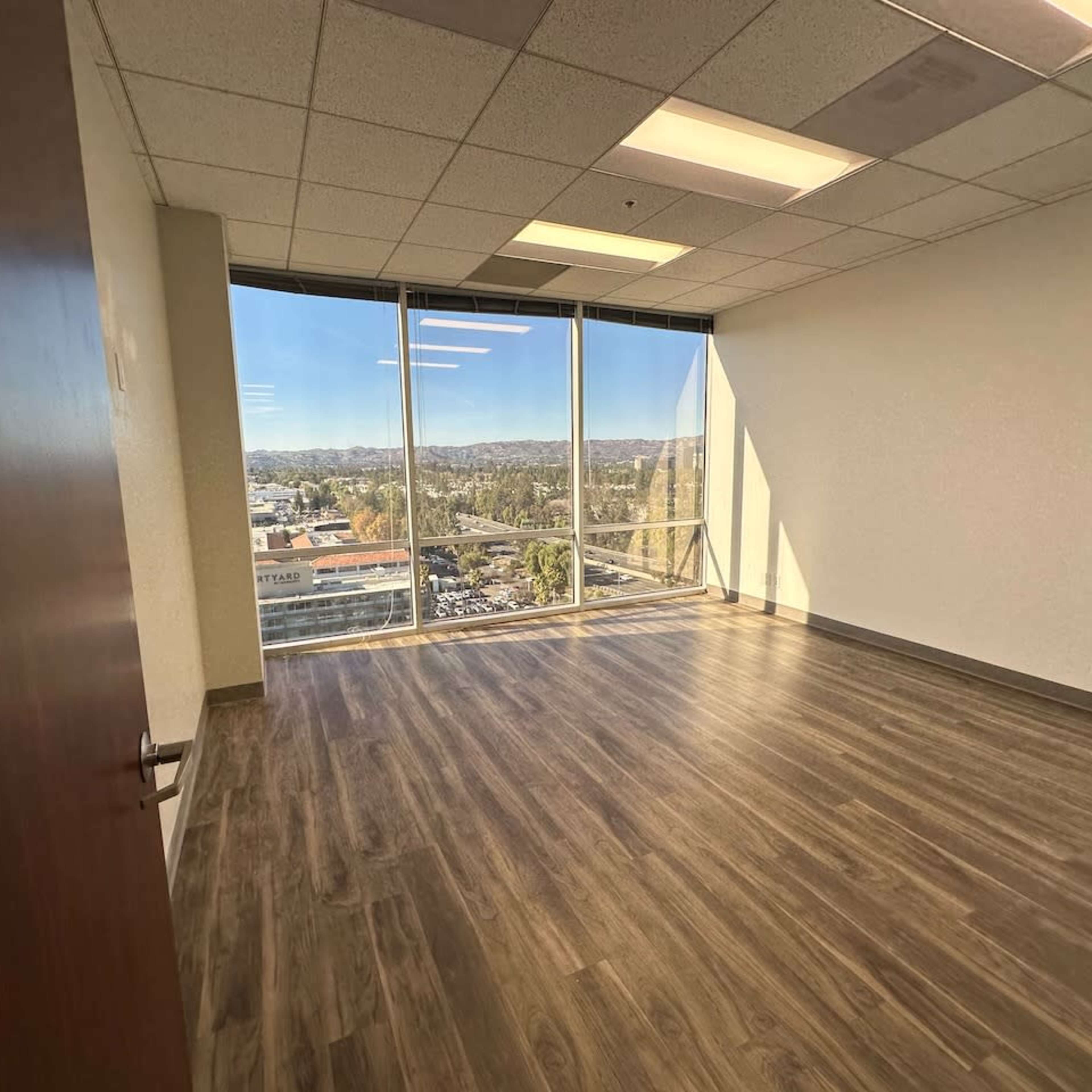 An empty office space with large windows offering a view of a parking lot and distant hills.