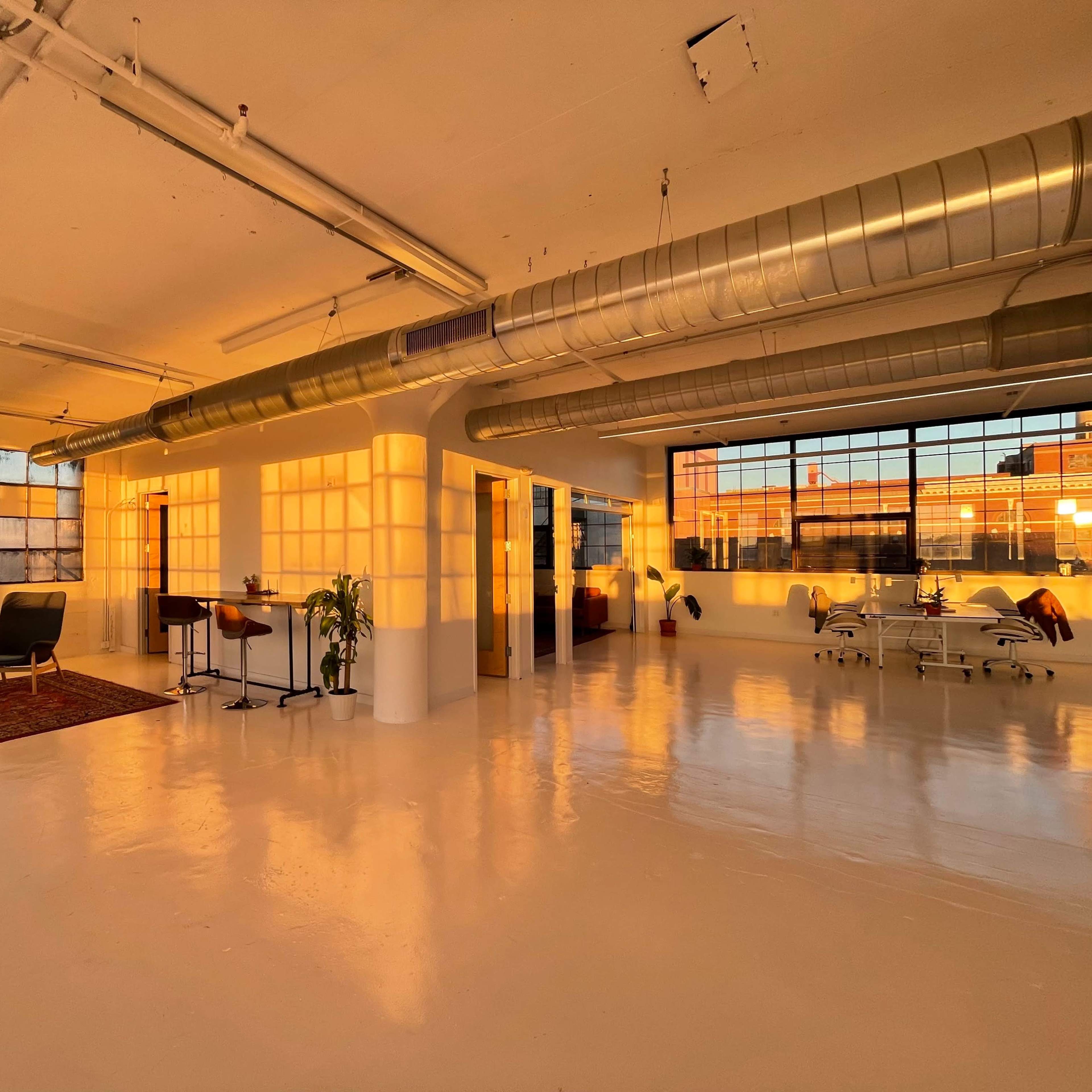 A spacious, well-lit industrial studio features large windows, polished white floors, and minimalist furnishings under soft sunset lighting.