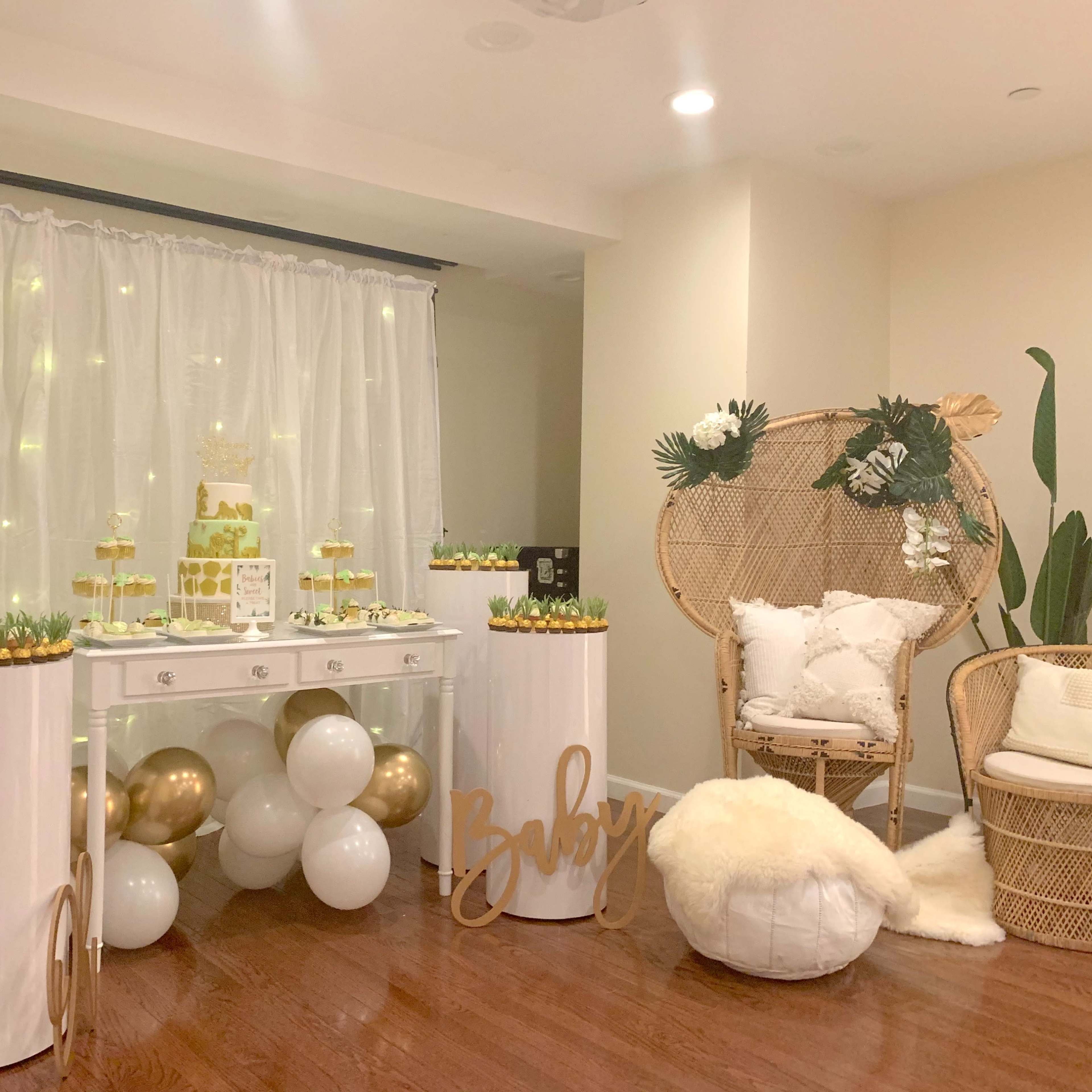 A decorated event space featuring a dessert table with treats displayed on a white table, surrounded by decorative accents, including gold balloons and a cozy seating area with a chair and a plush throw.