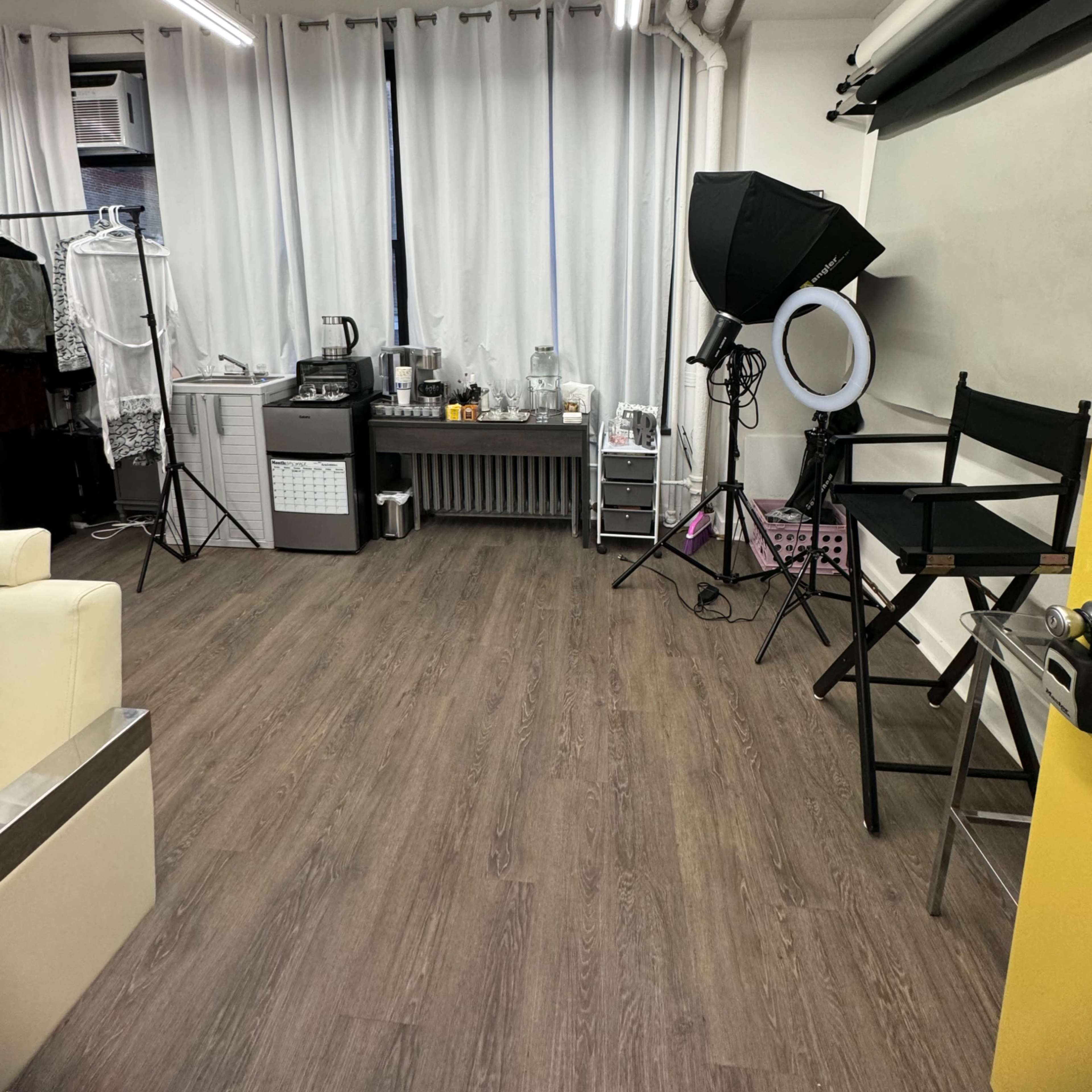 A spacious room with a wooden floor, featuring white curtains, a kitchenette area, a lighting setup, and a black director's chair.