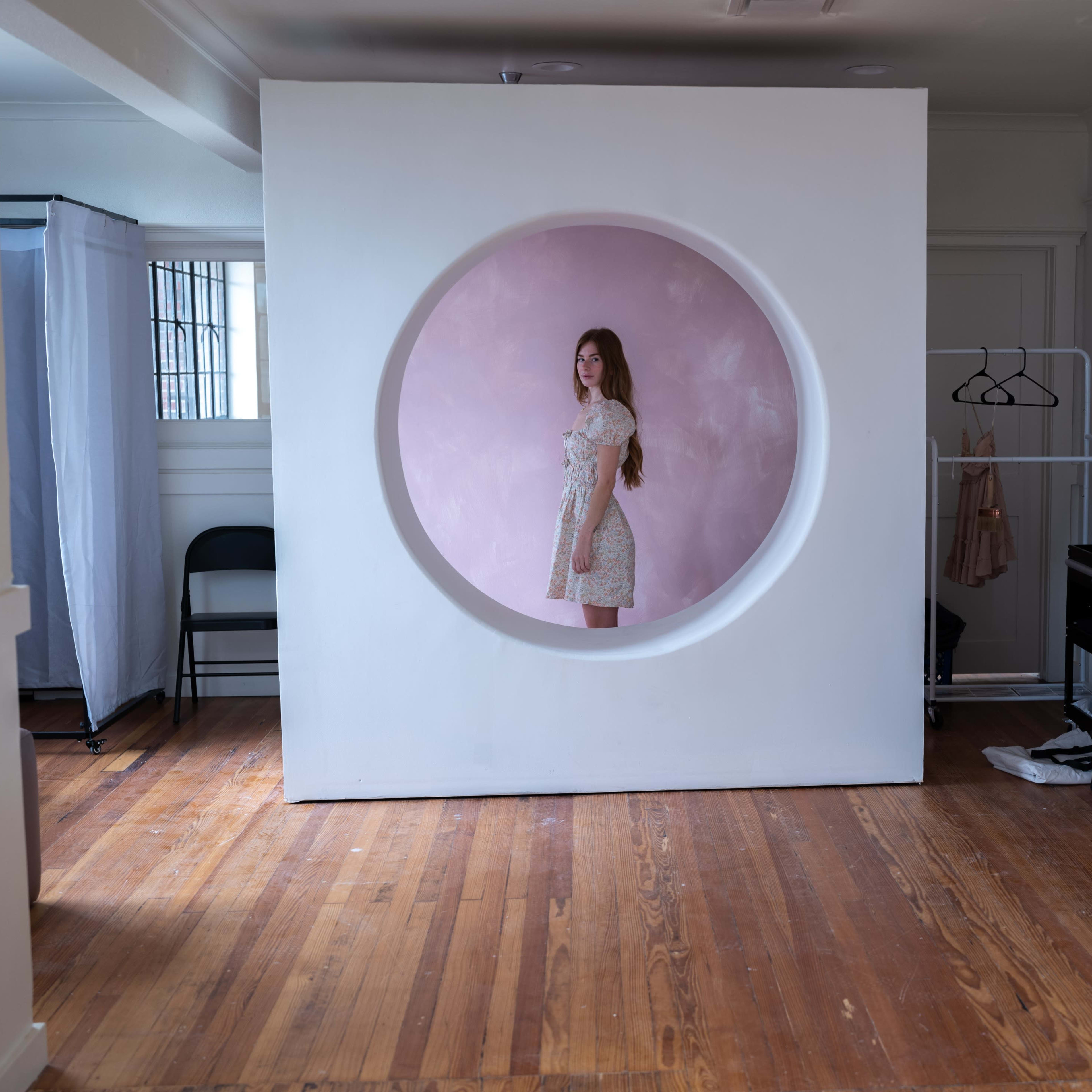 A young woman stands inside a circular cutout in a white wall, set against a pink backdrop in a room with wooden floors and minimal furnishings.