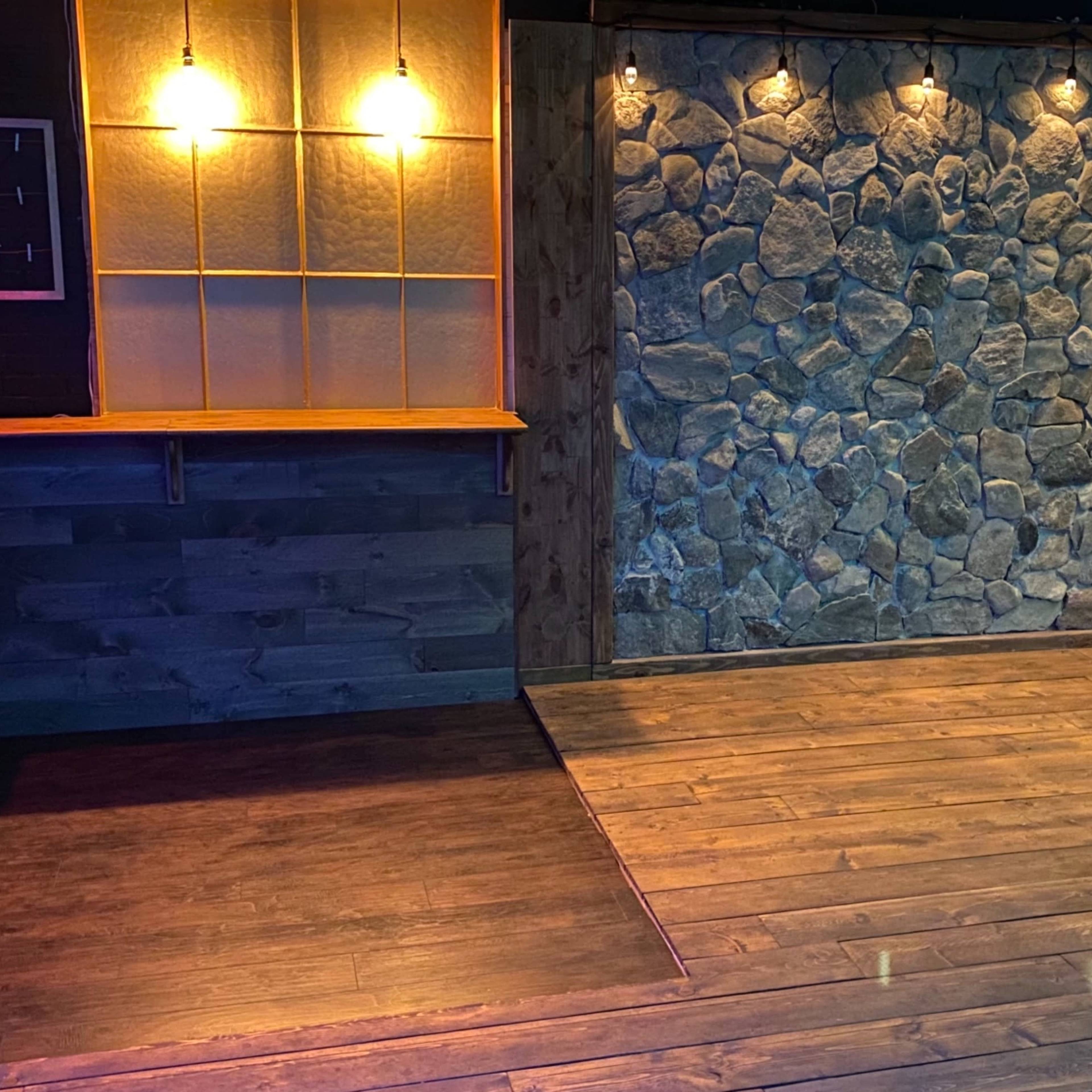 A cozy indoor space featuring a wooden floor, a stone wall, and warm lighting, with a bar area on the left and greenery on the right.