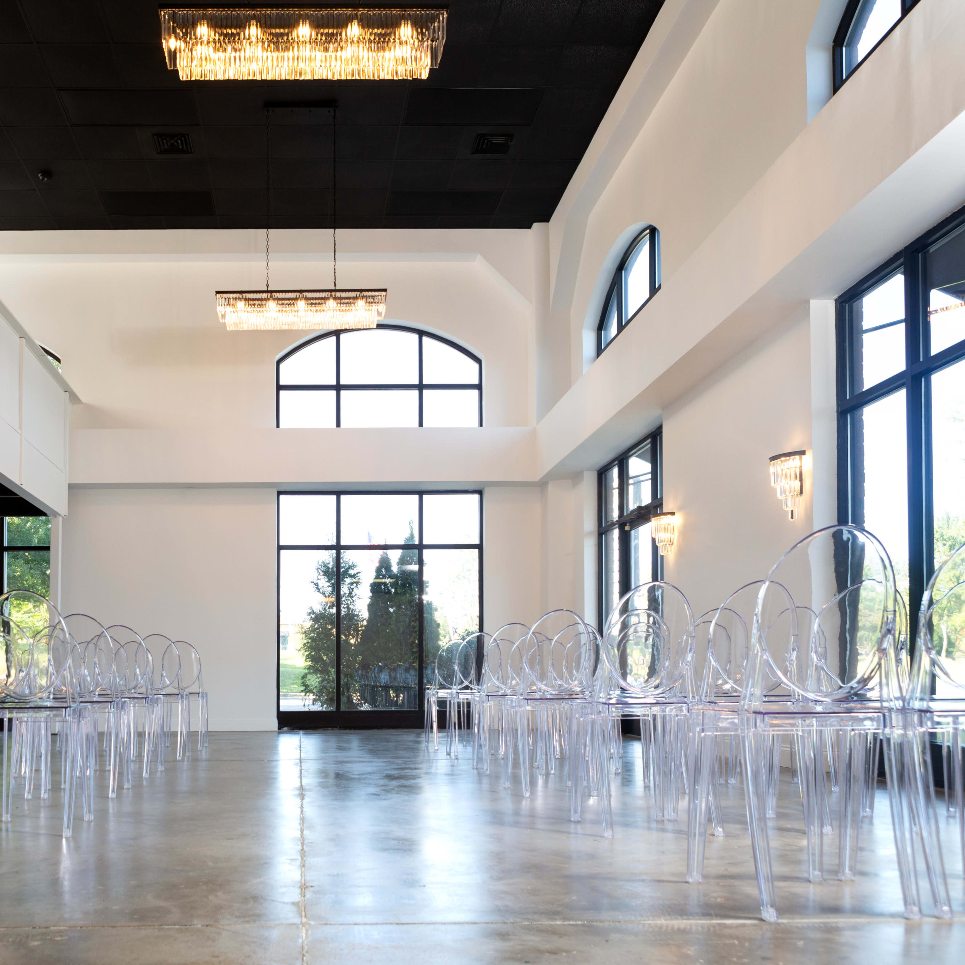 A spacious, modern indoor venue with clear acrylic chairs arranged in rows, large windows allowing natural light, and elegant light fixtures on the walls.