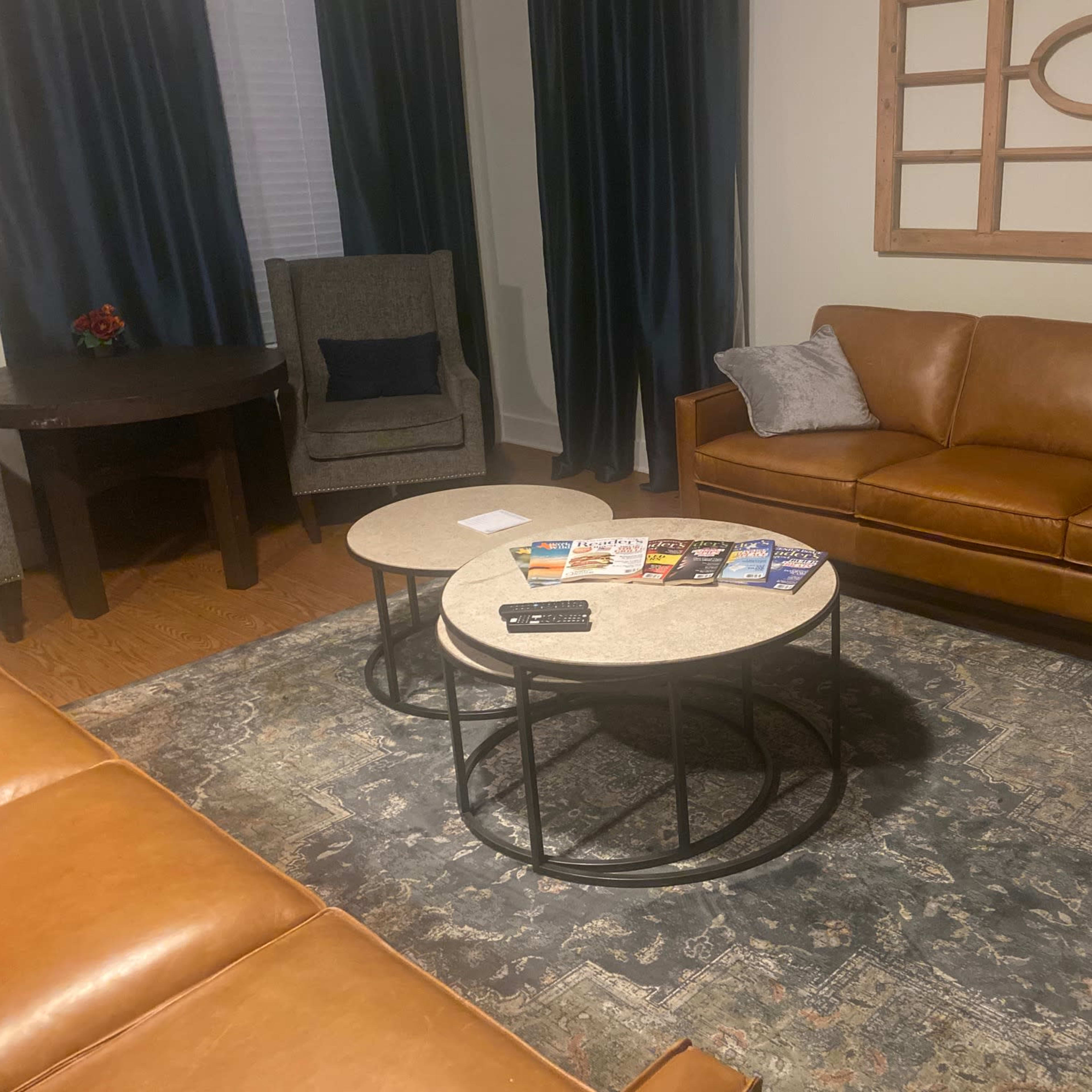 A living room with a brown leather sofa, two coffee tables with magazines arranged on top, and a patterned rug.