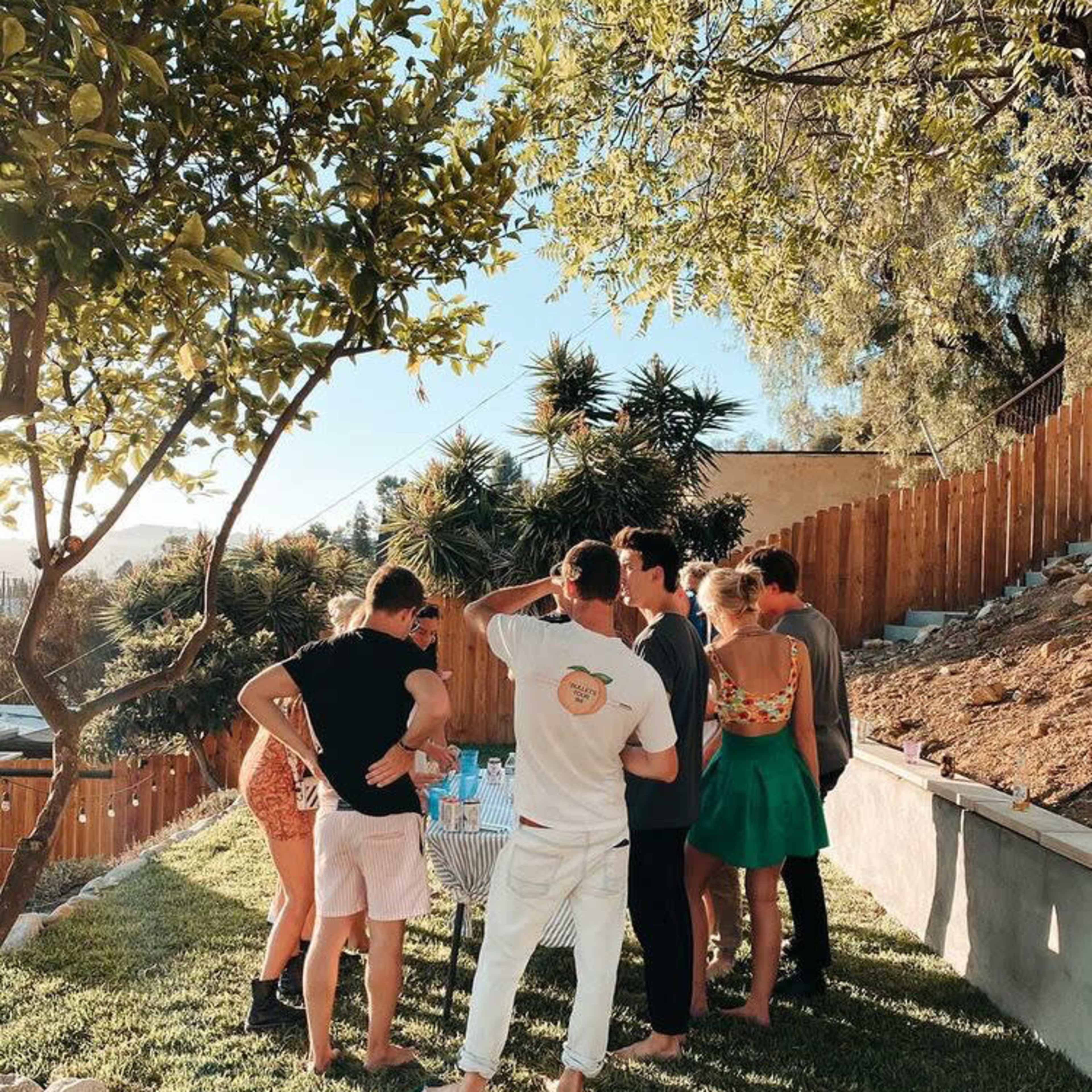 A group of seven people gathers around a table in a backyard, with a mix of greenery and wooden fencing in the background.