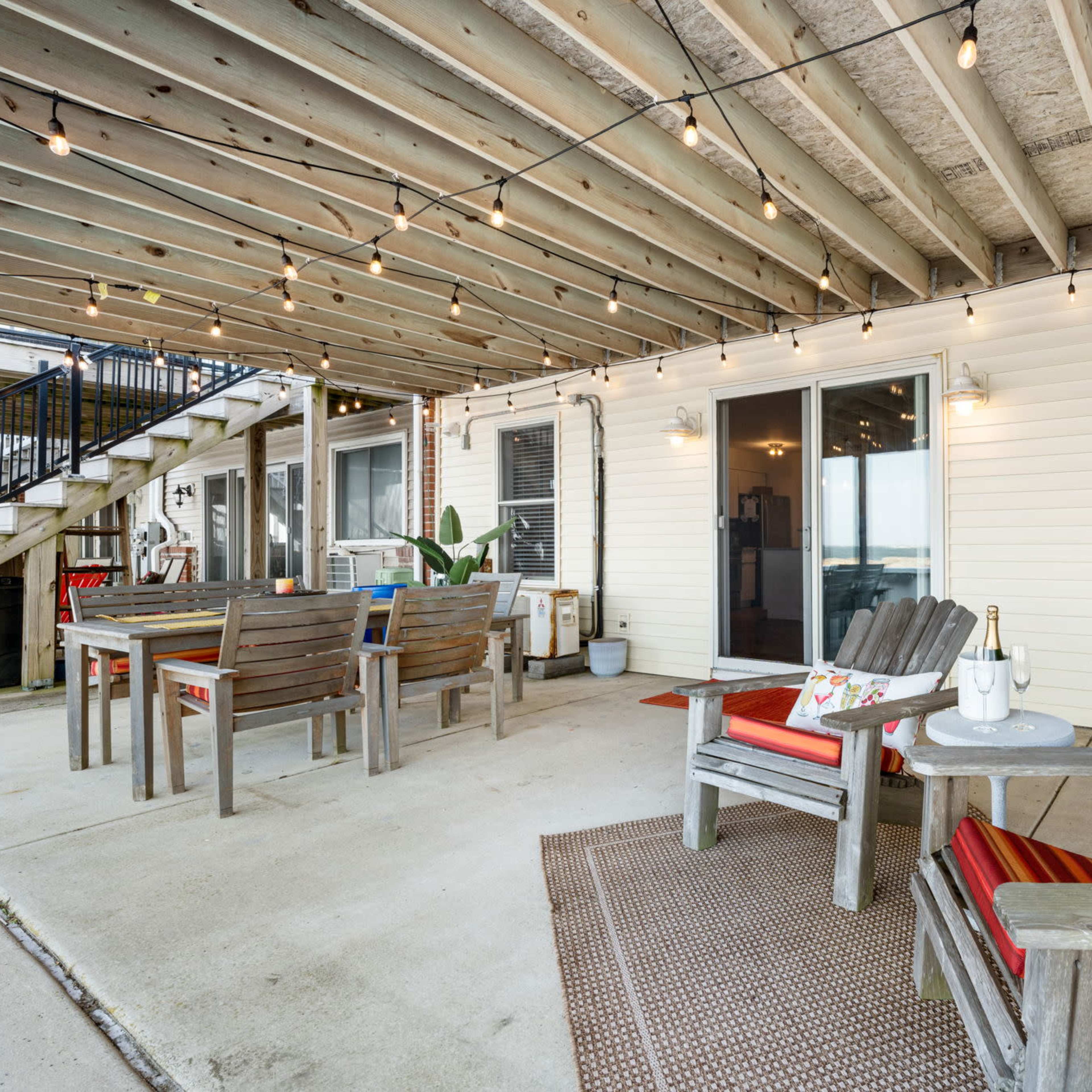 A covered patio area with wooden furniture, string lights overhead, and a small table with a bottle of champagne and a glass.