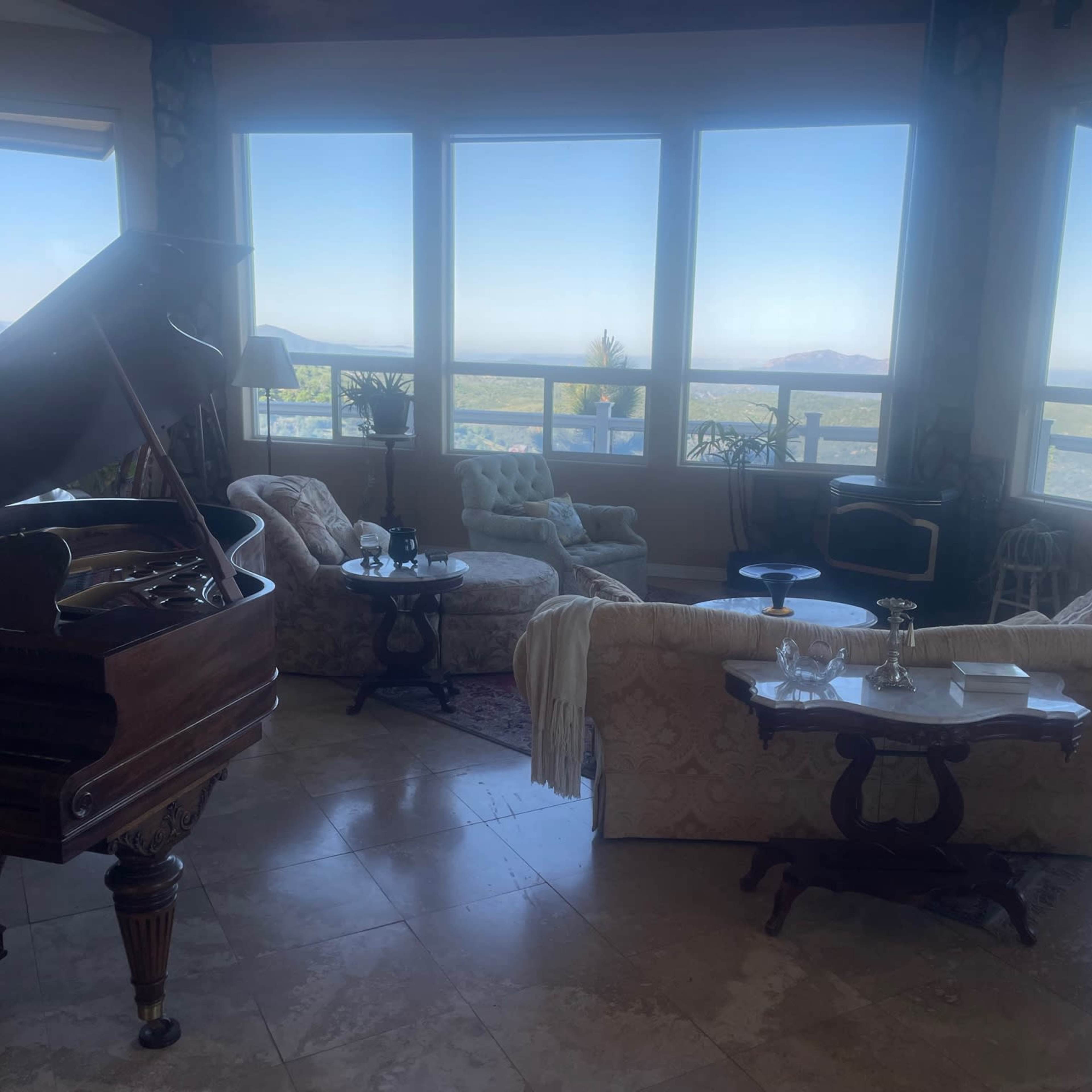 A spacious living room with large windows overlooking a landscape, furnished with a grand piano, several upholstered chairs, and a coffee table.