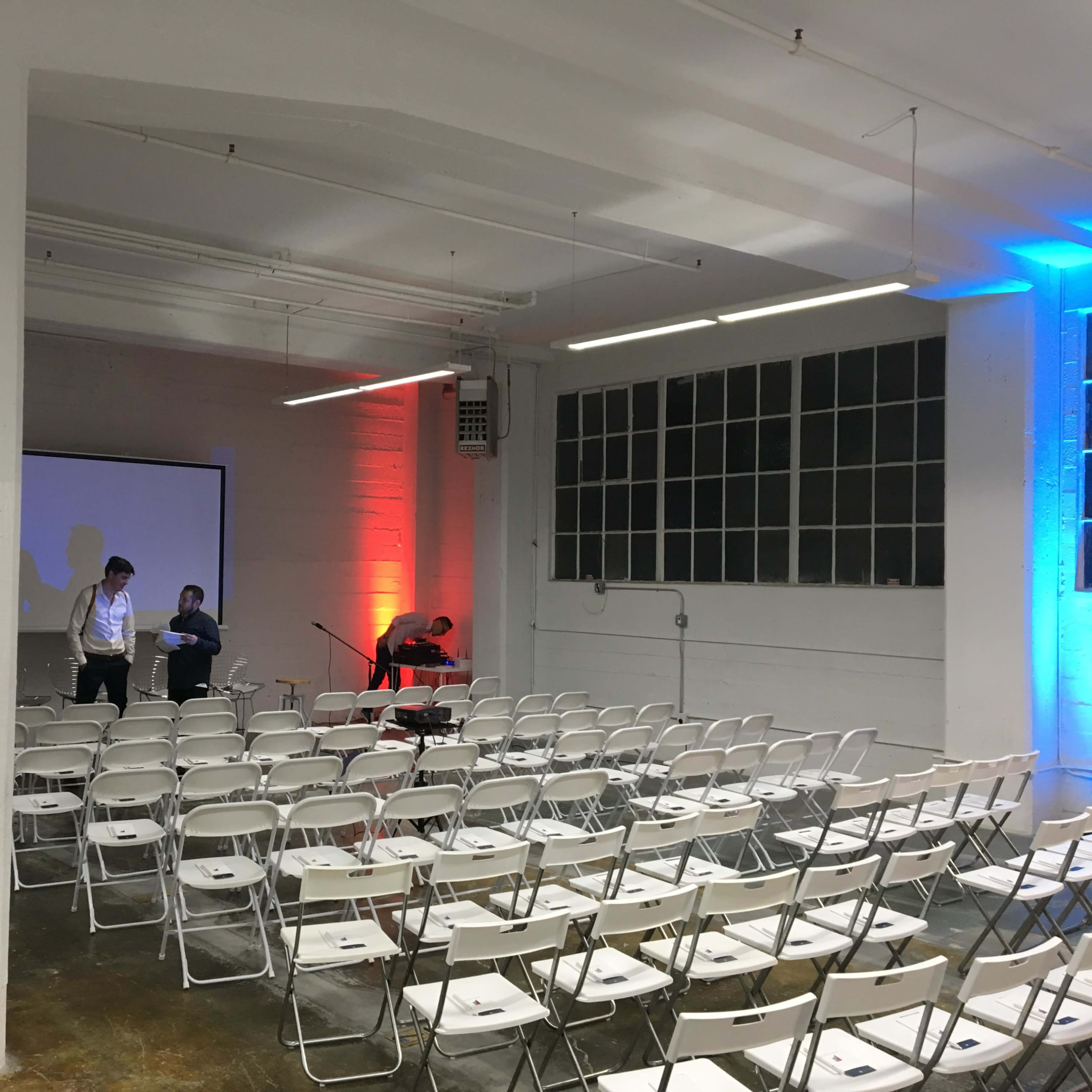 A spacious room with rows of white folding chairs set up for an event, and two people preparing a presentation on a stage with a large screen.