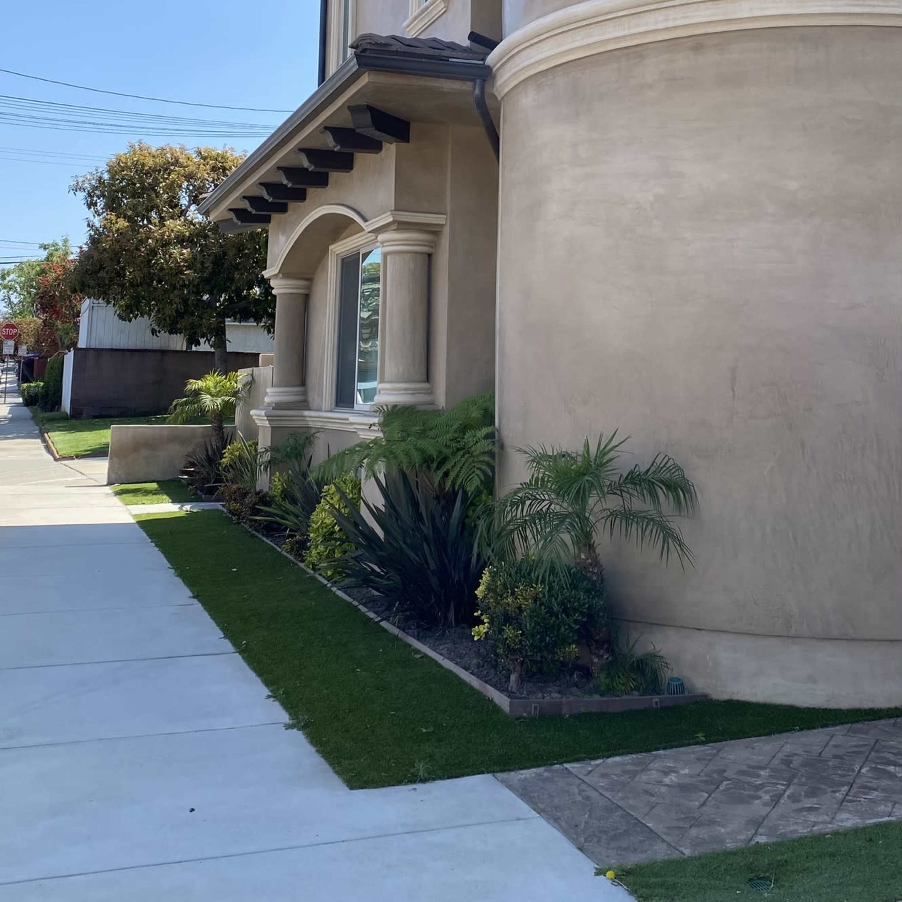 A two-story house with a curved, beige exterior and large windows is situated on a sidewalk lined with greenery.