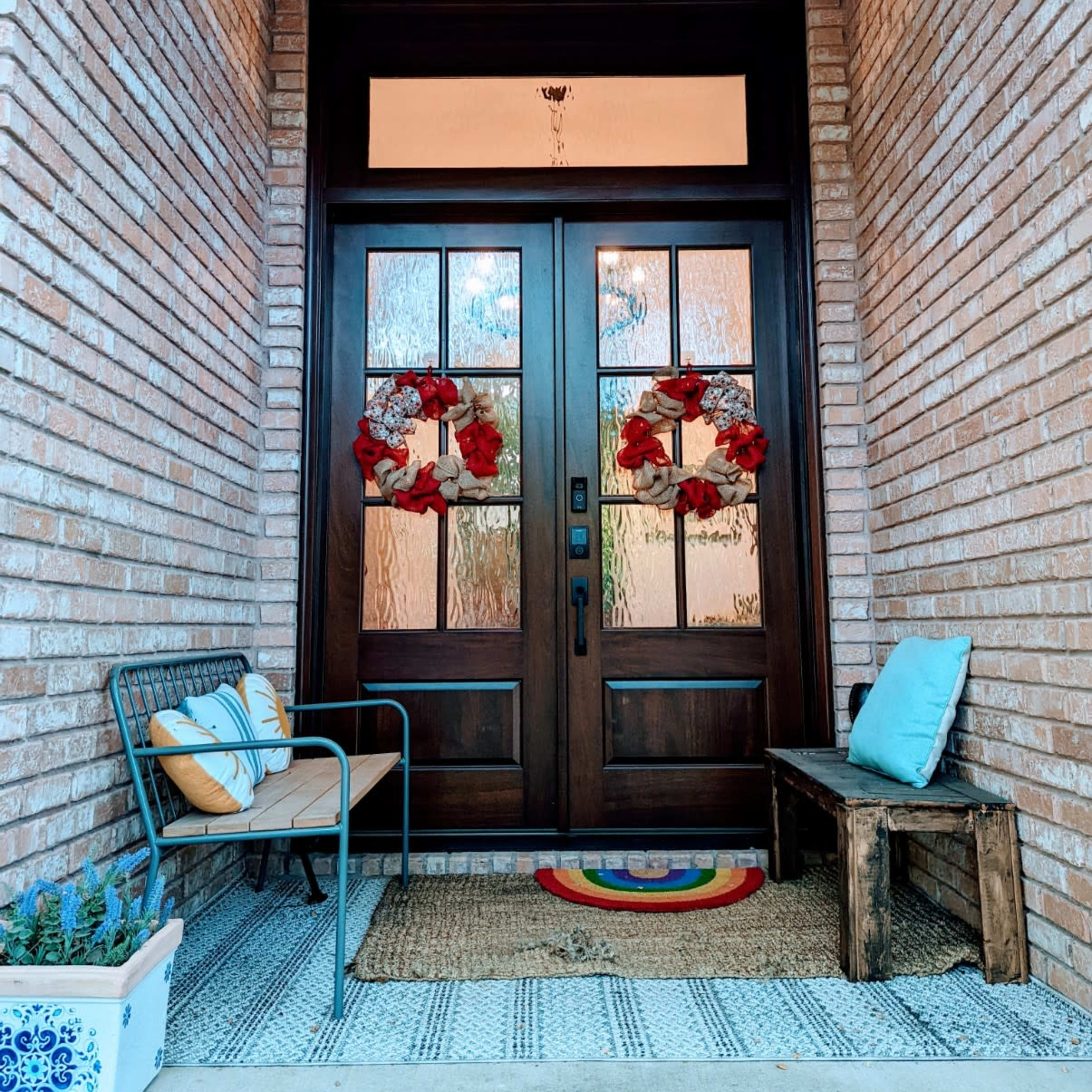 A welcoming entrance featuring a pair of wooden double doors with glass panels, adorned with red wreaths, flanked by a bench and chair on a textured mat.