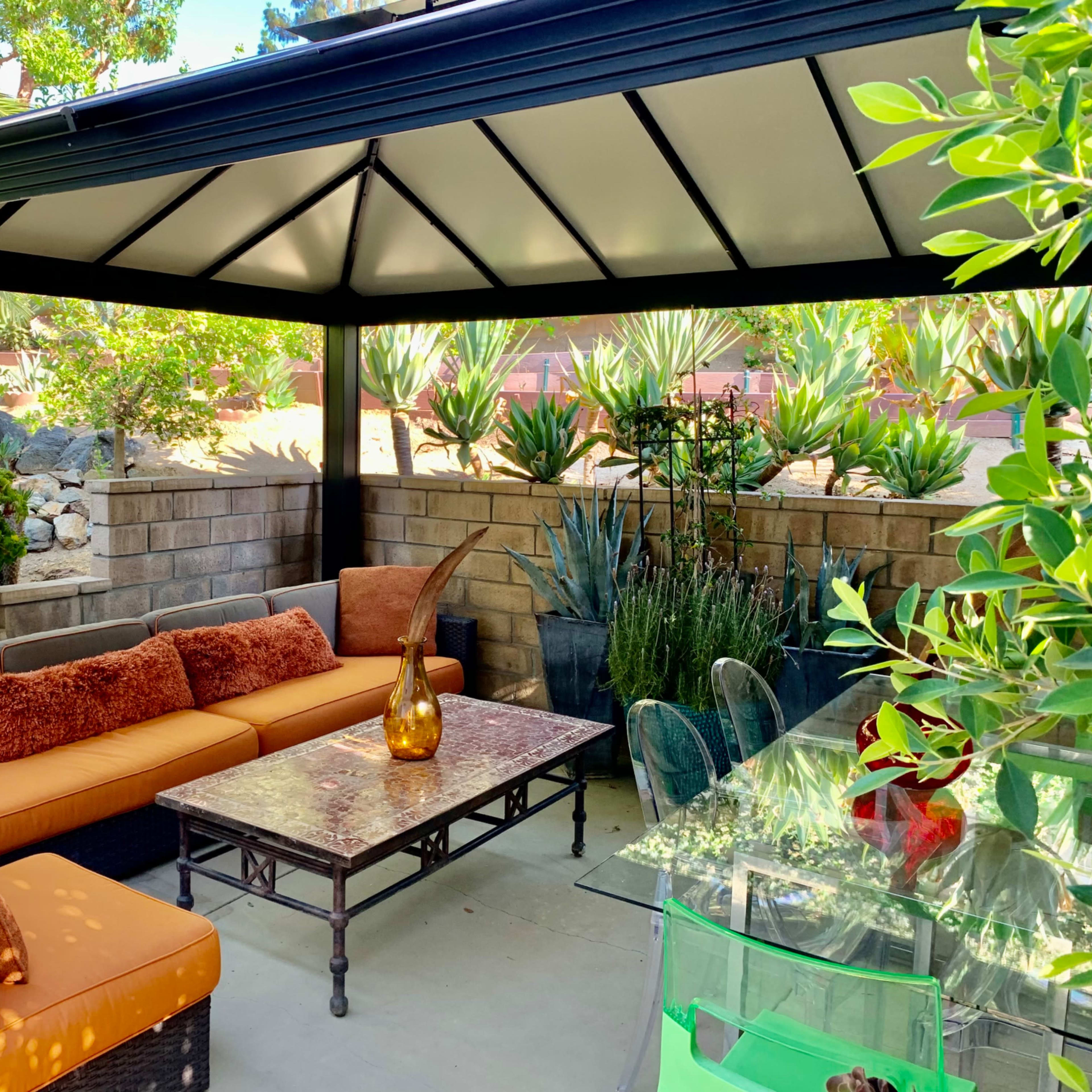A shaded outdoor seating area with a sofa, a glass table, and various potted plants around a stone wall.