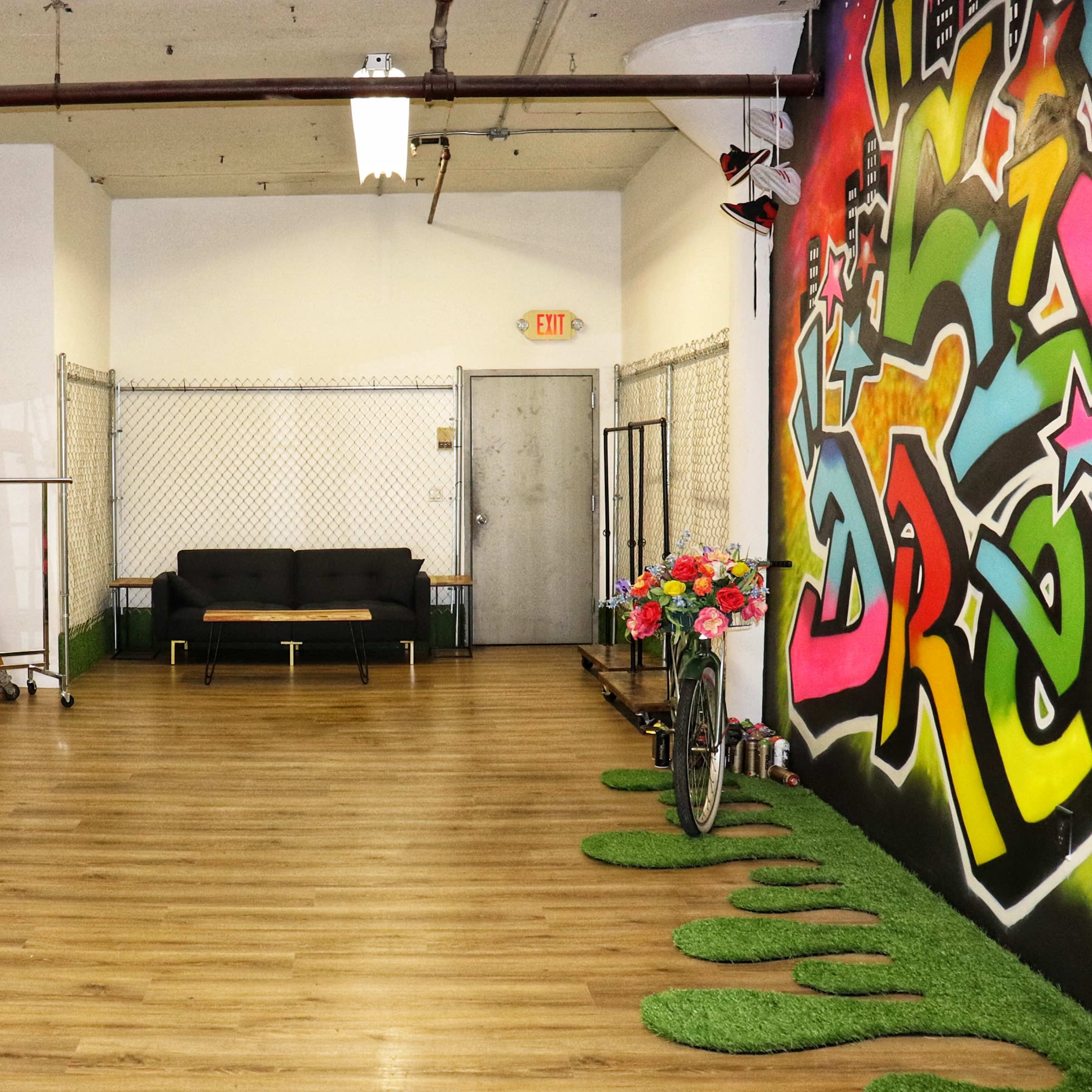 A spacious interior with wooden flooring, a black couch, a ladder leaning against the wall, and a colorful graffiti mural on one side.