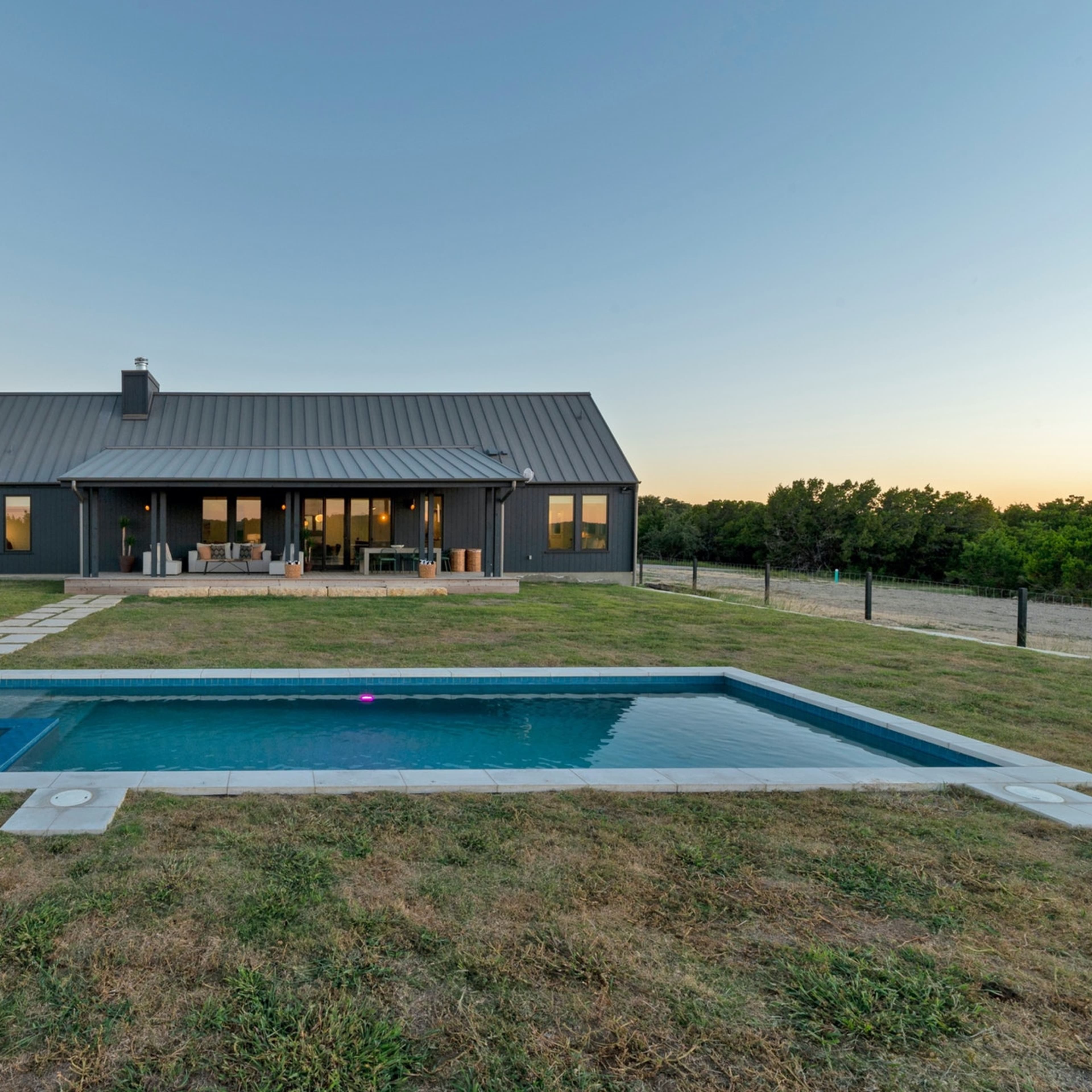 A modern house with a metal roof is located beside a rectangular swimming pool and surrounded by a grassy yard and trees at dusk.