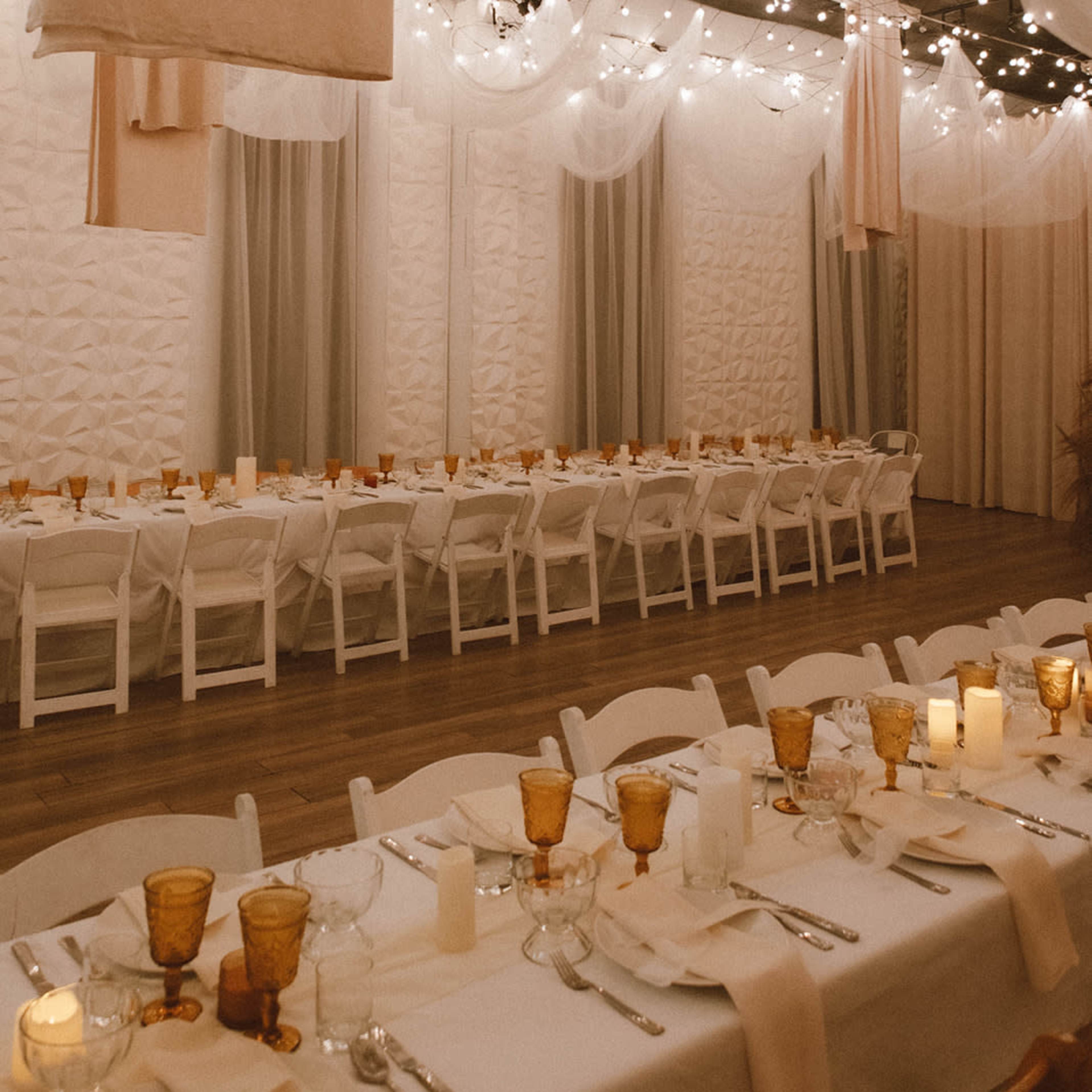 A banquet hall features long tables set with glasses and candles, beneath draped fabric and string lights.