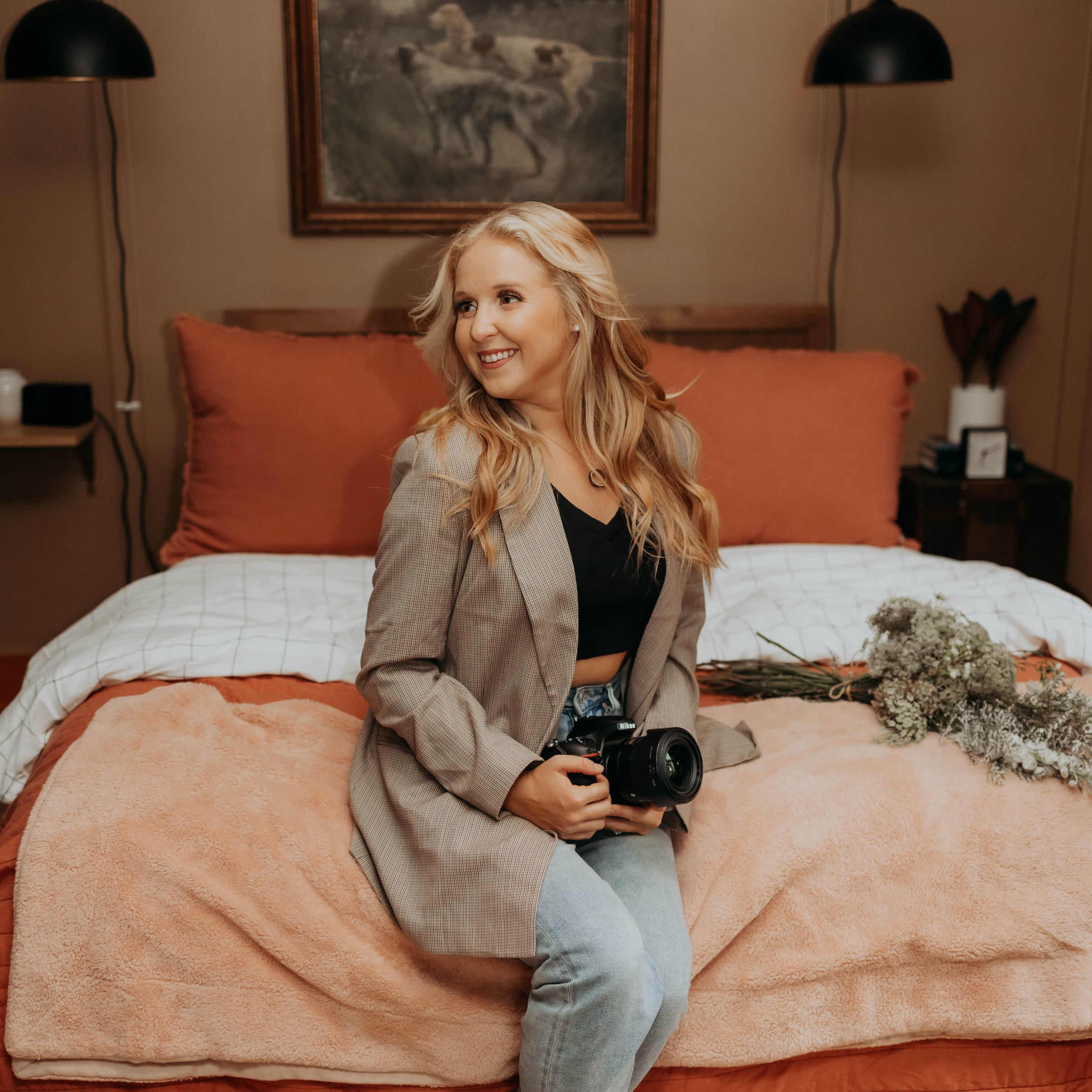 A woman sits on a bed holding a camera, dressed in a blazer and jeans, with decorative plants and a painting in the background.