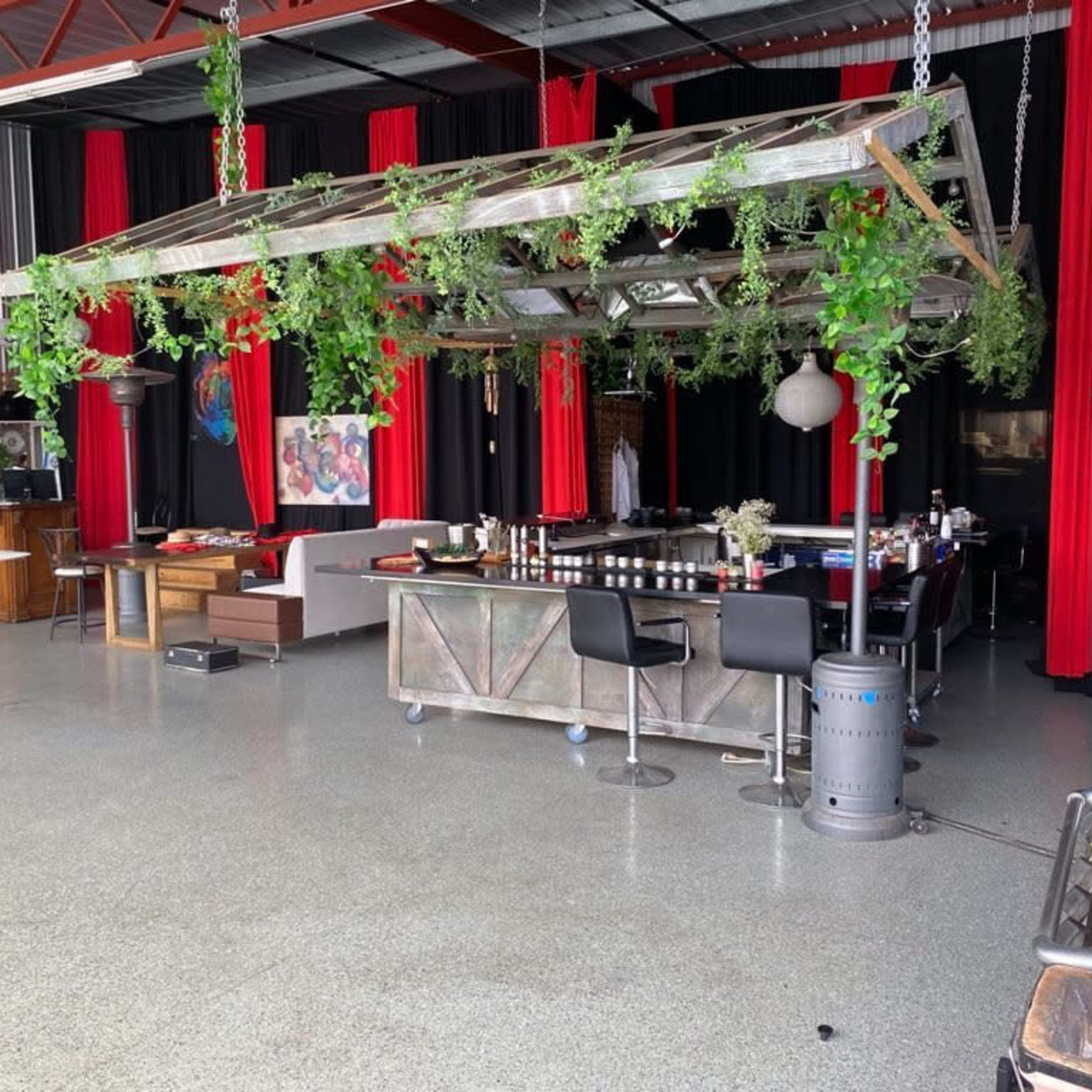 A spacious indoor venue features a bar surrounded by greenery and red curtains, with various seating areas and artistic decor.