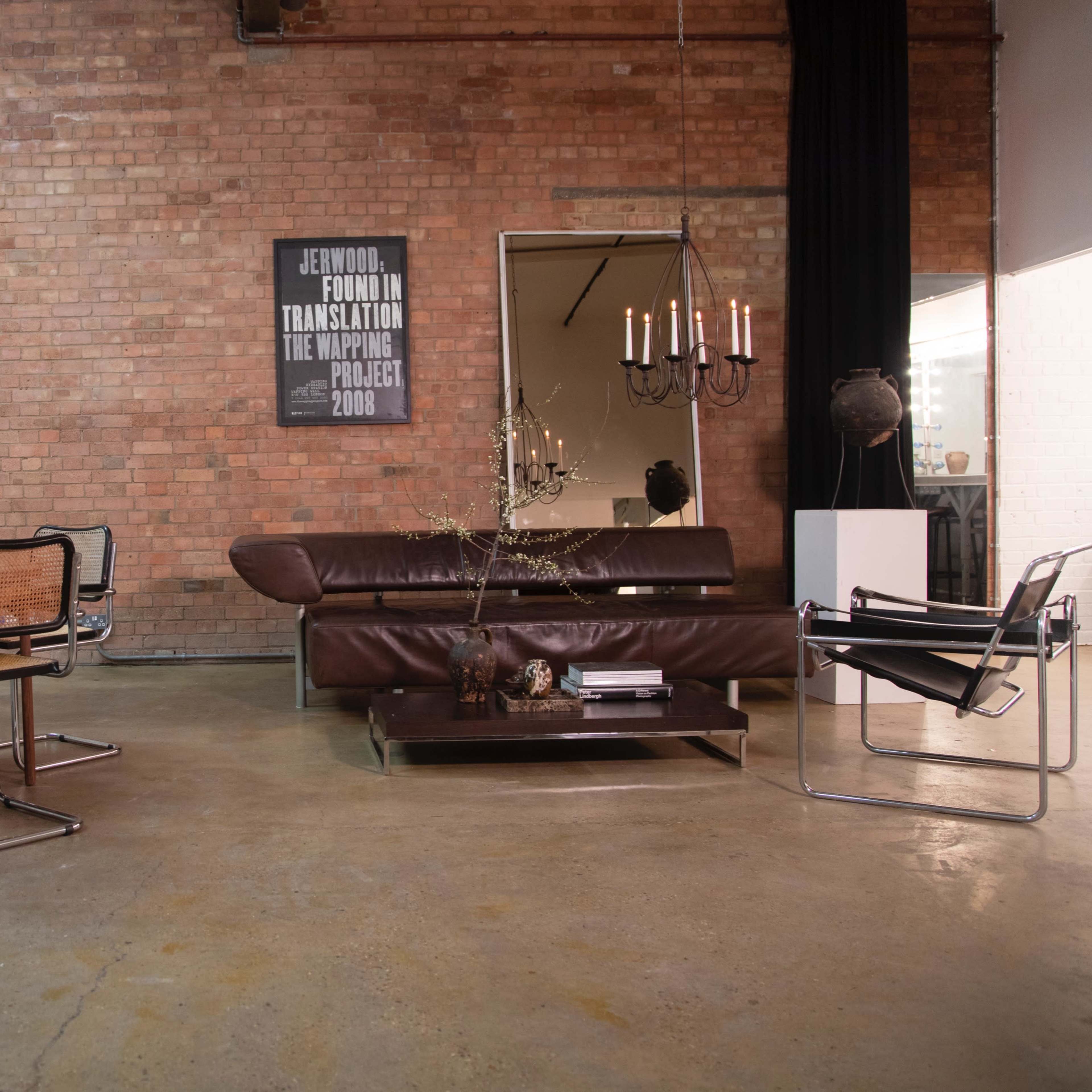The scene features a minimalist living space with a round wooden dining table, four black and chrome chairs, a brown leather sofa, a large vase with flowers, and a wall-mounted artwork in a room with exposed brick walls.