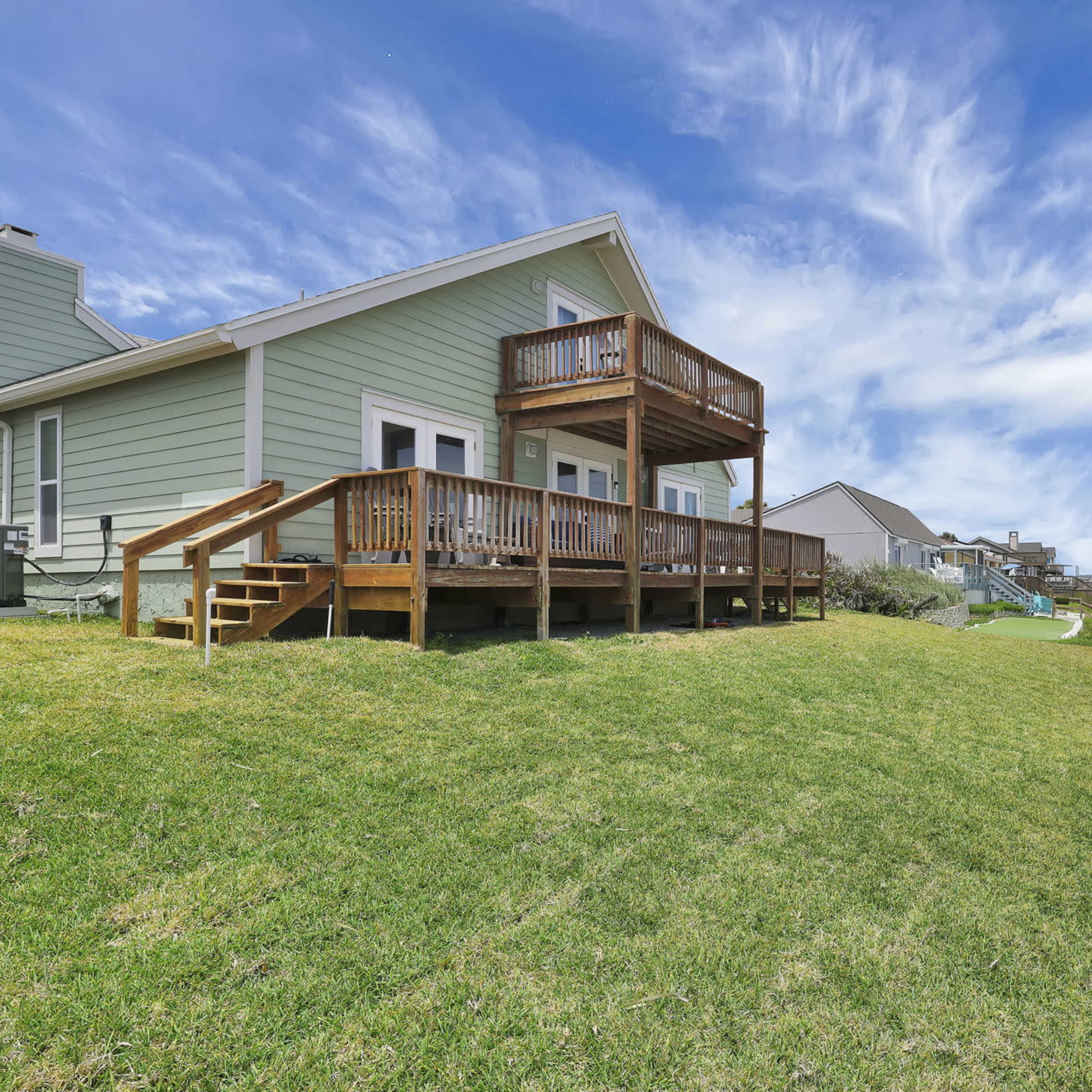A beach house with a wooden deck overlooks the sandy shore under a partly cloudy sky.